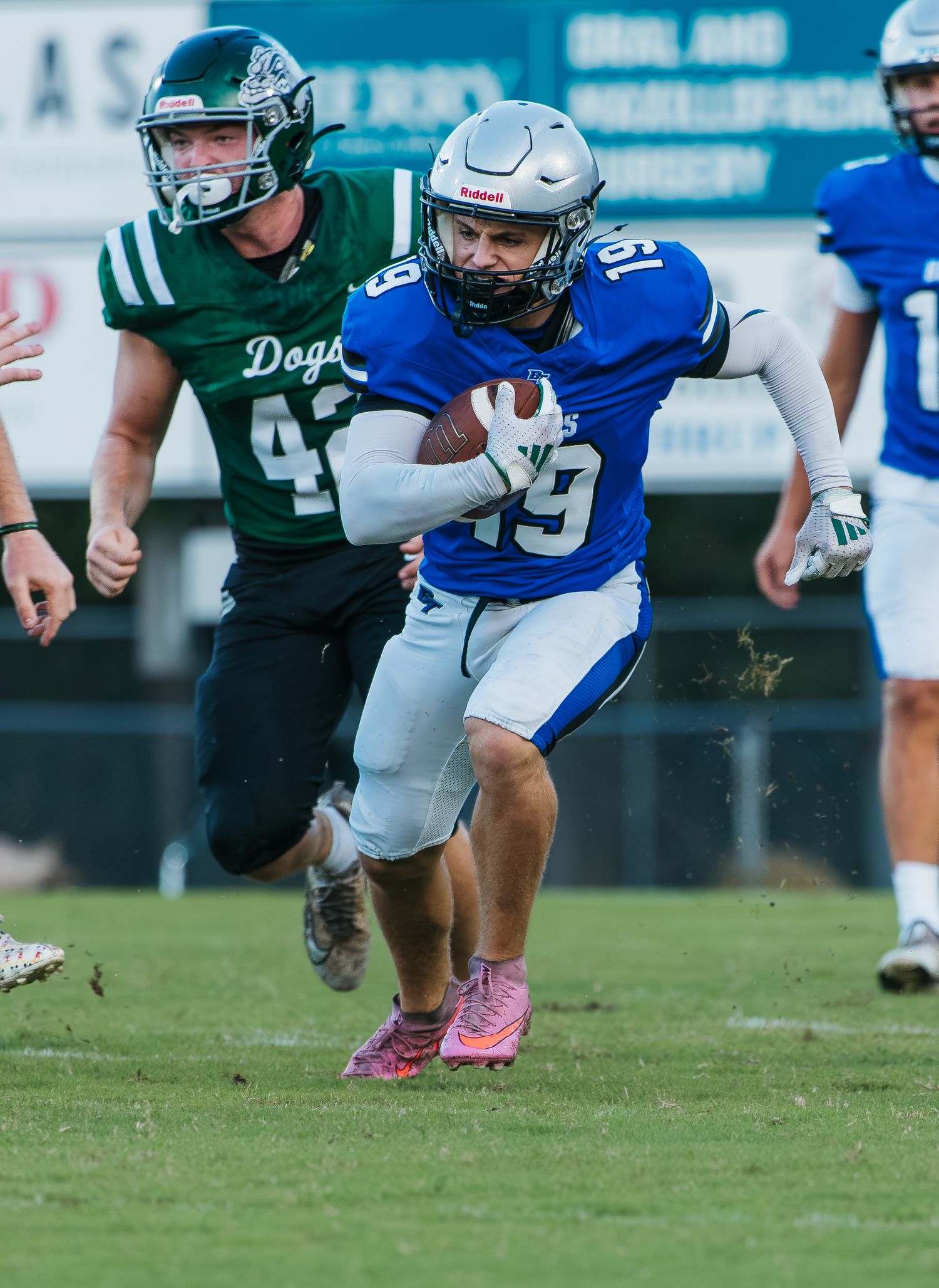 High school football player in blue jersey #19 runs with ball while green-jersied defender #42 pursues during game.