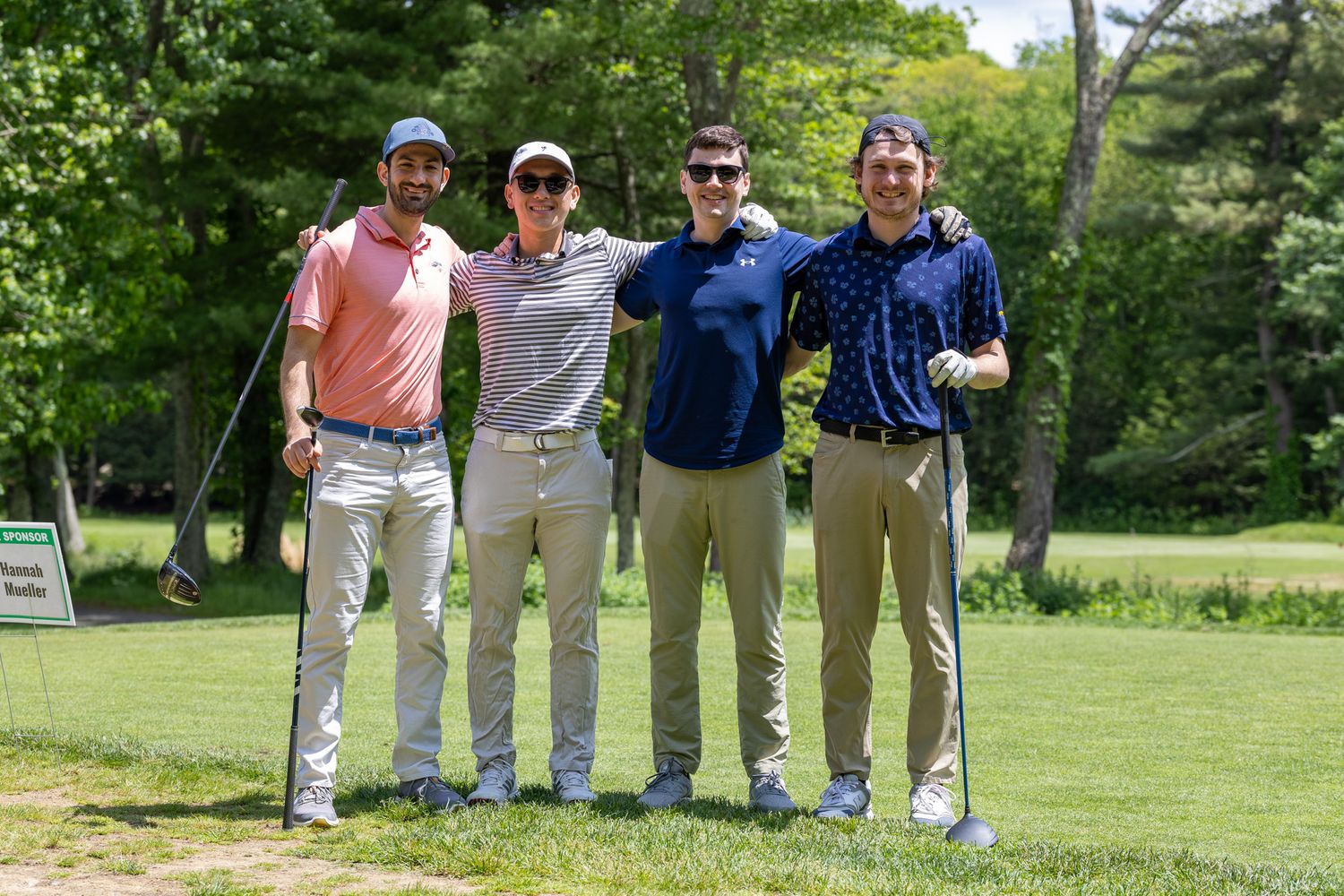 Four men pose with golf clubs on a sunny golf course surrounded by lush green trees.