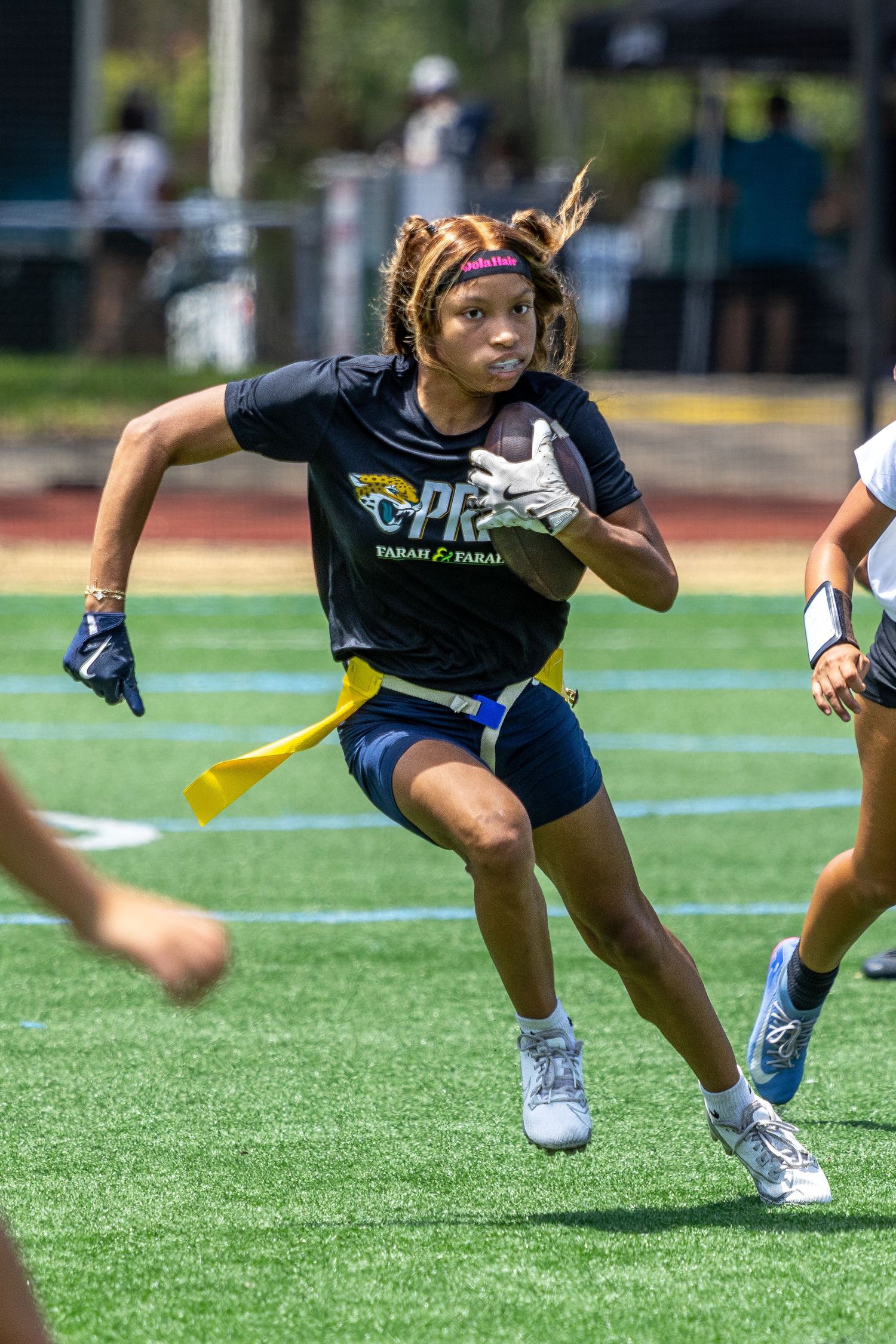 Young female athlete runs with football during flag football game on turf field, wearing black jersey and blue shorts.