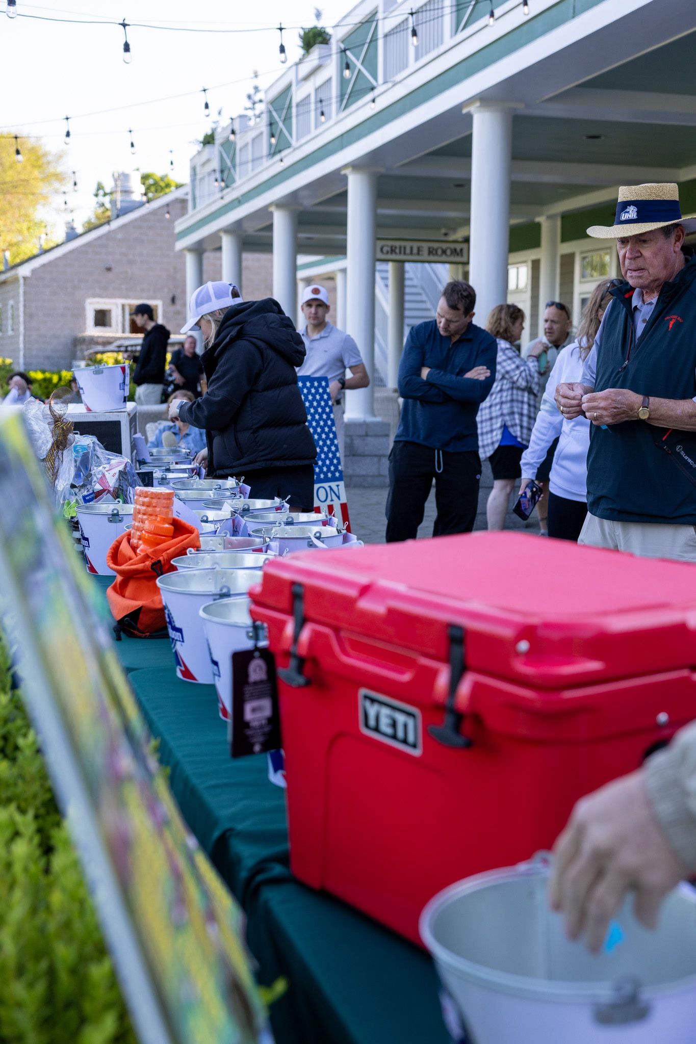 Outdoor charity auction event with a red YETI cooler and prize table, guests browsing items near a clubhouse.