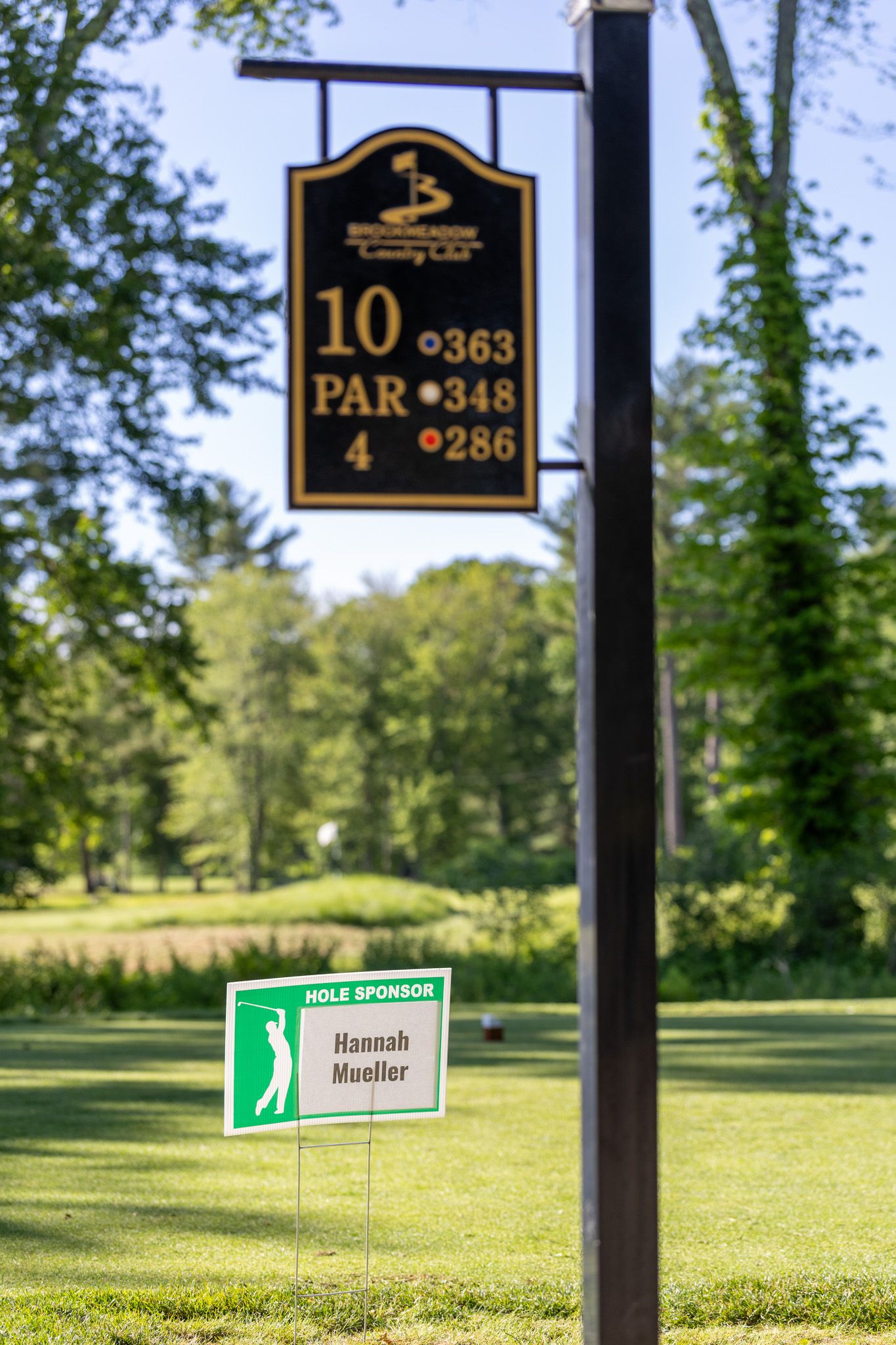 Golf course hole 10 sign showing par 4 with yardages, beside a Hannah Mueller hole sponsor sign on a sunny day.