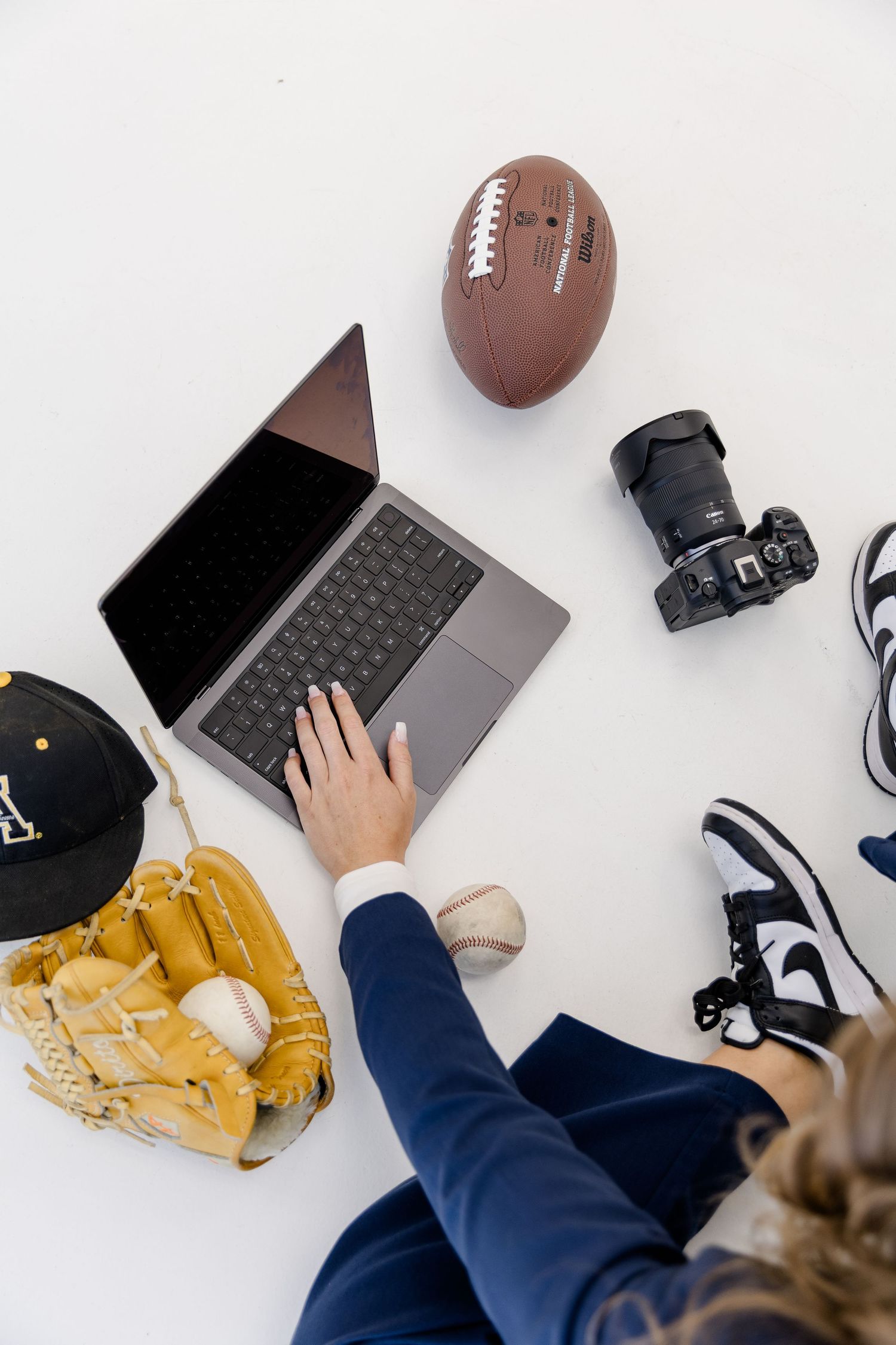 Person using laptop surrounded by sports equipment including football, baseball glove, camera, and Nike sneakers on white background.