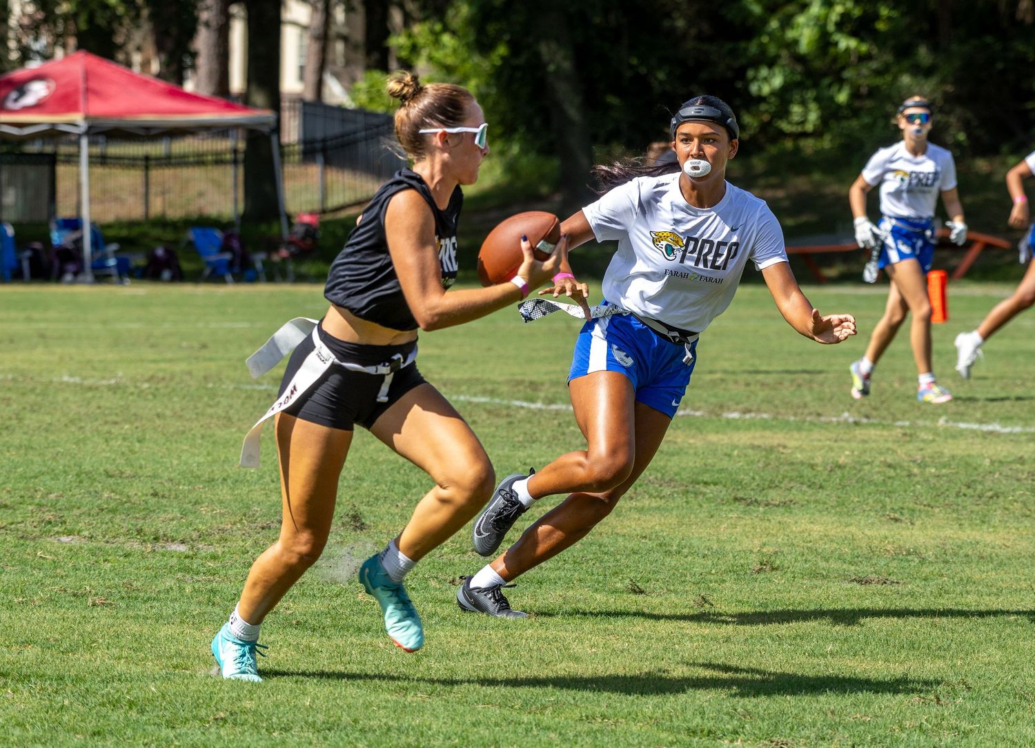 Two female athletes compete in a flag football game on a sunny grass field, one catching a pass while the other defends.