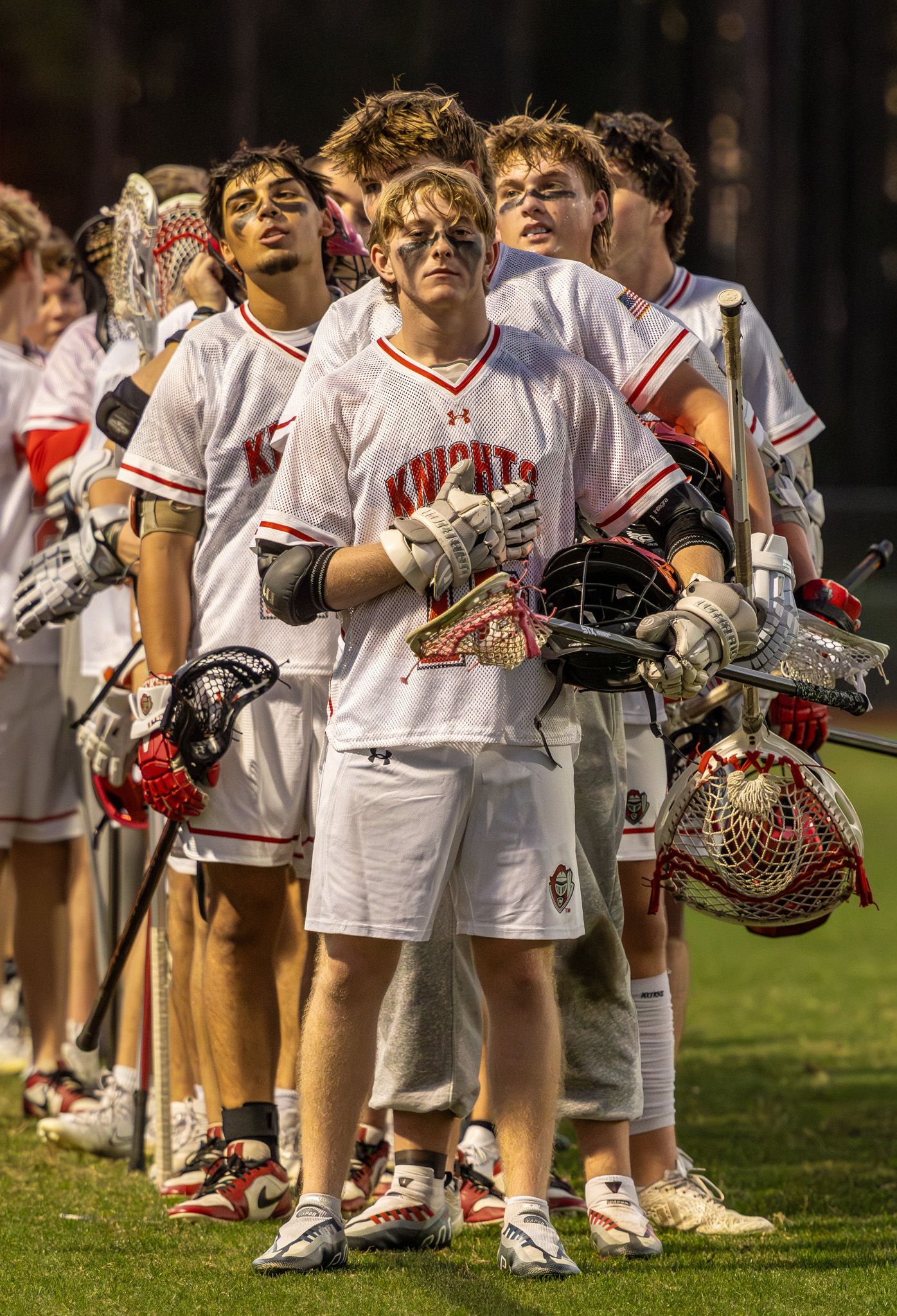 Knights lacrosse team players in white uniforms holding helmets and sticks stand together on field during game.