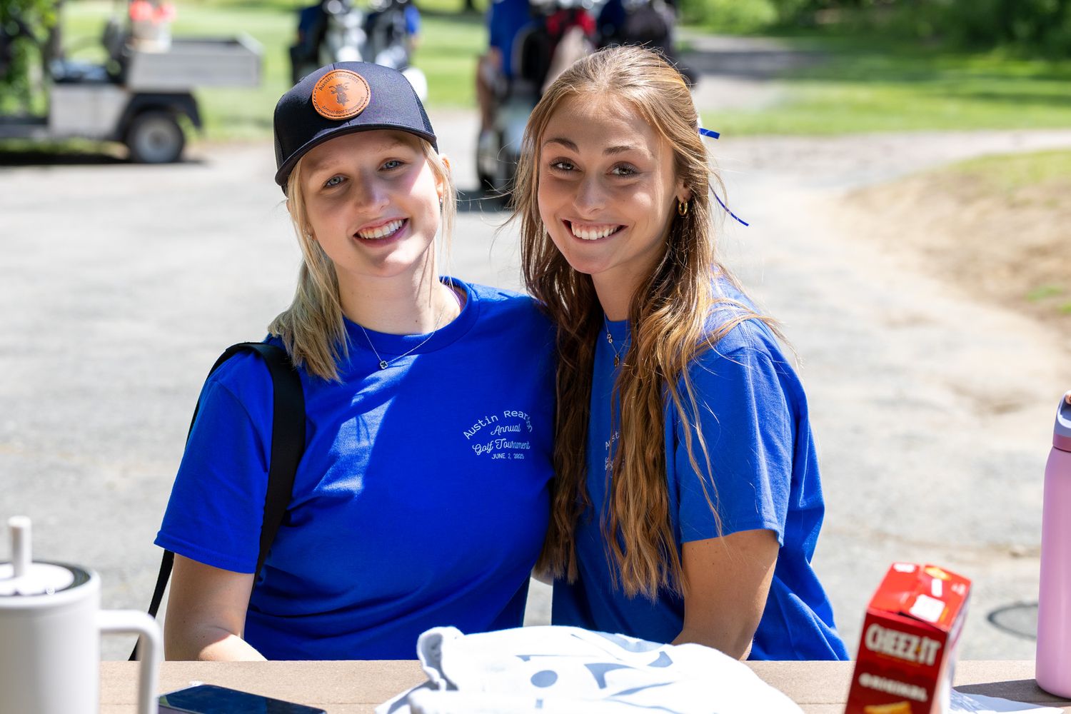 Two smiling women in blue t-shirts pose together outdoors at a sunny event with Cheez-It box visible.