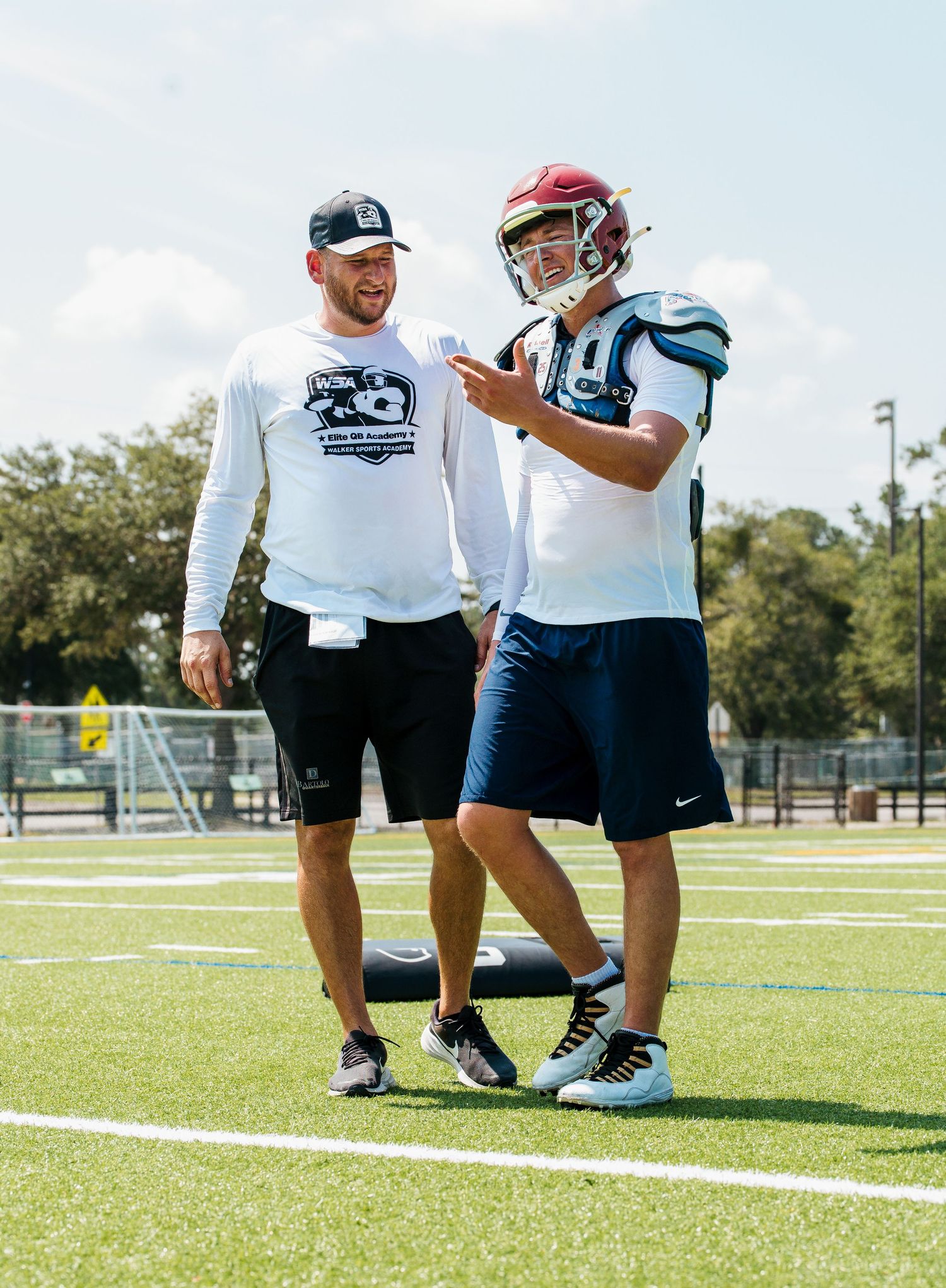 Two football players in uniform discuss plays on a green turf field during an outdoor practice session.