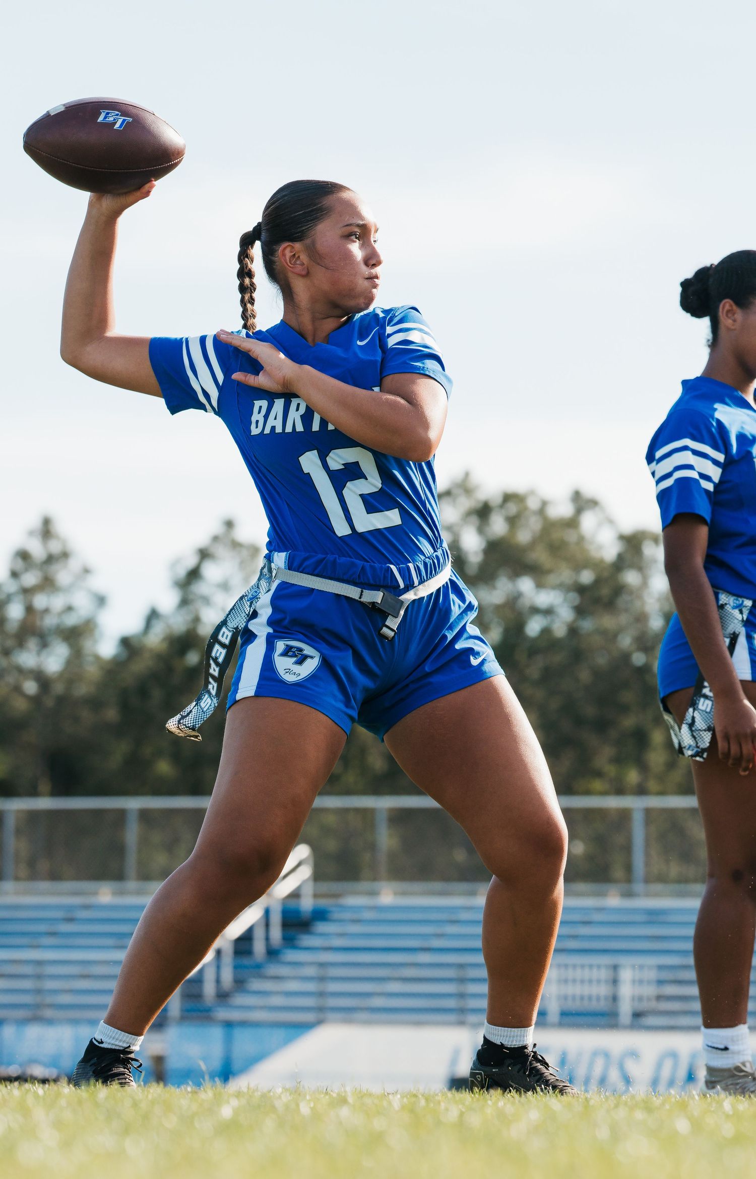 Female flag football player #12 in blue Bart jersey throws a pass during an outdoor game.