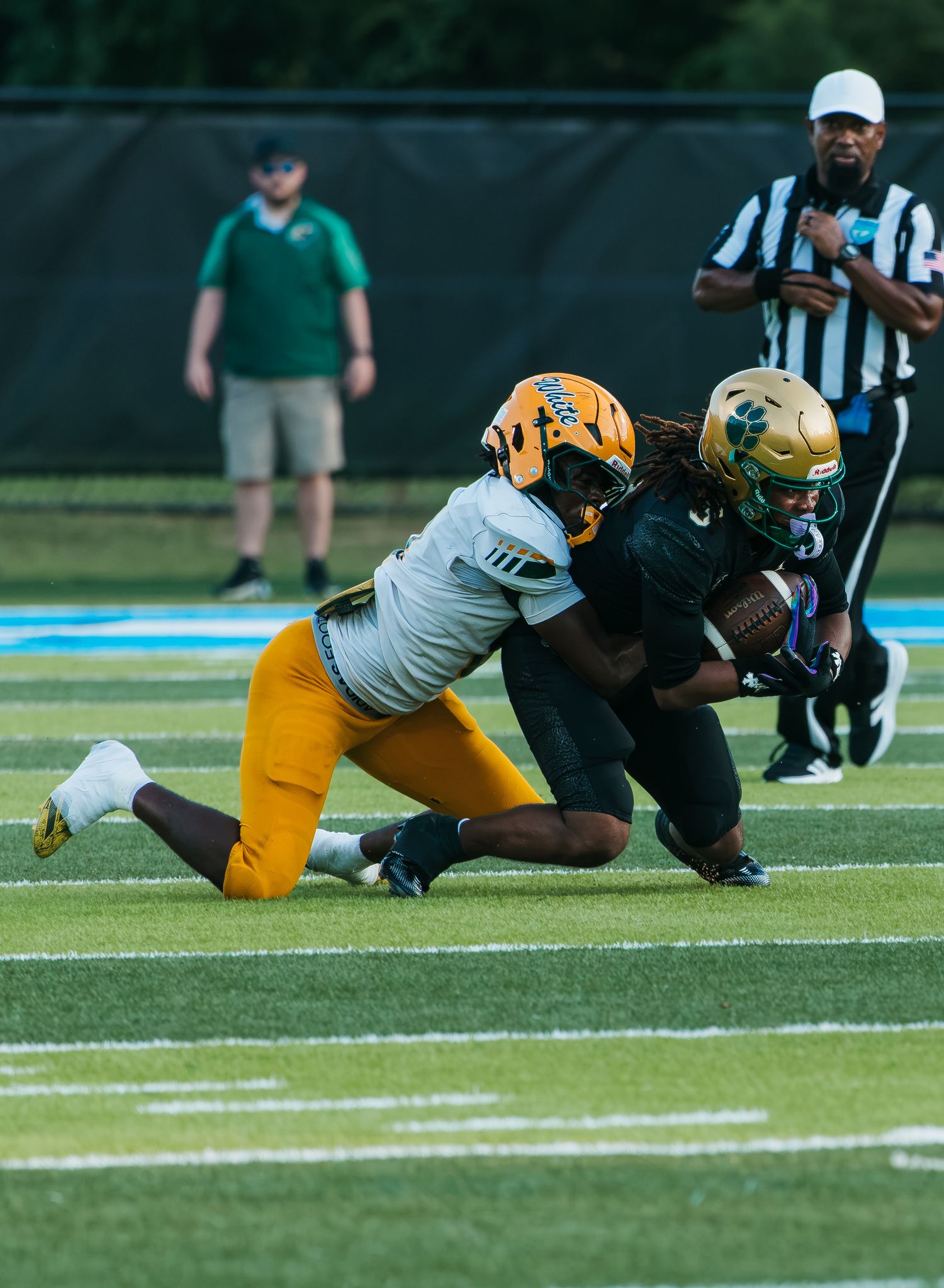 Football player in gold helmet tackles opponent in black uniform during a competitive game on a green turf field.