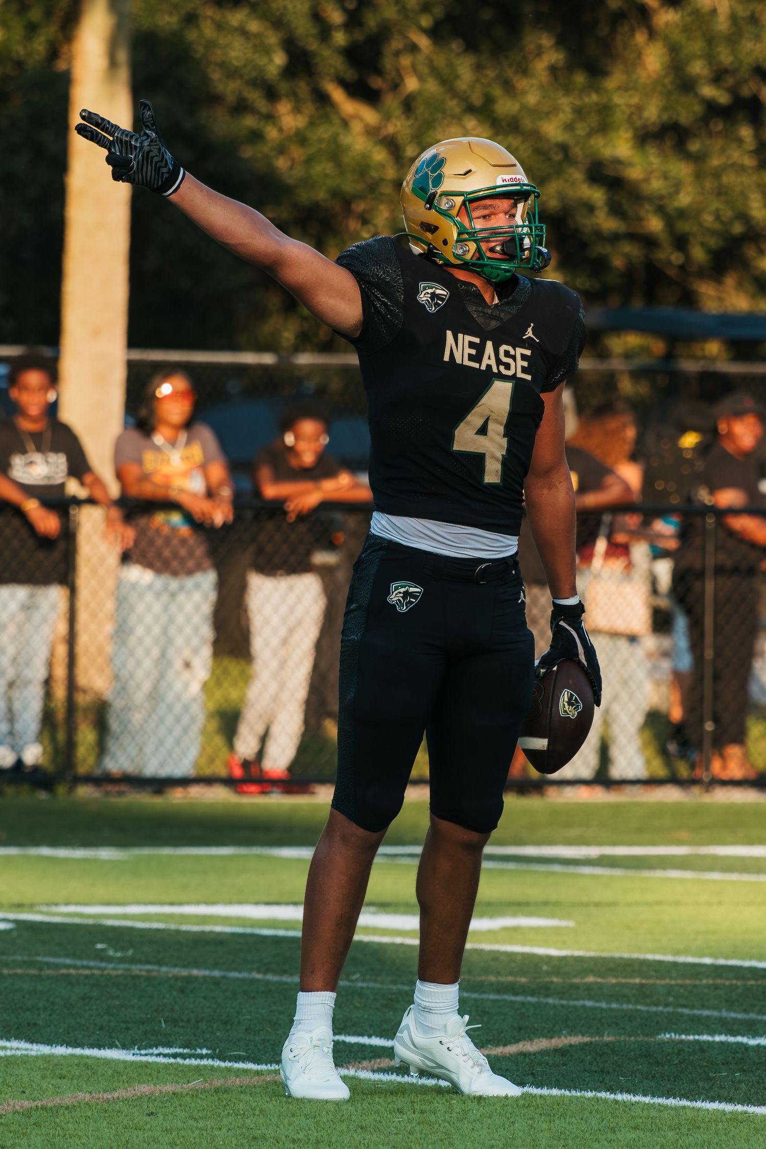 Nease football player #4 points skyward in black and gold uniform during a game under evening lights.
