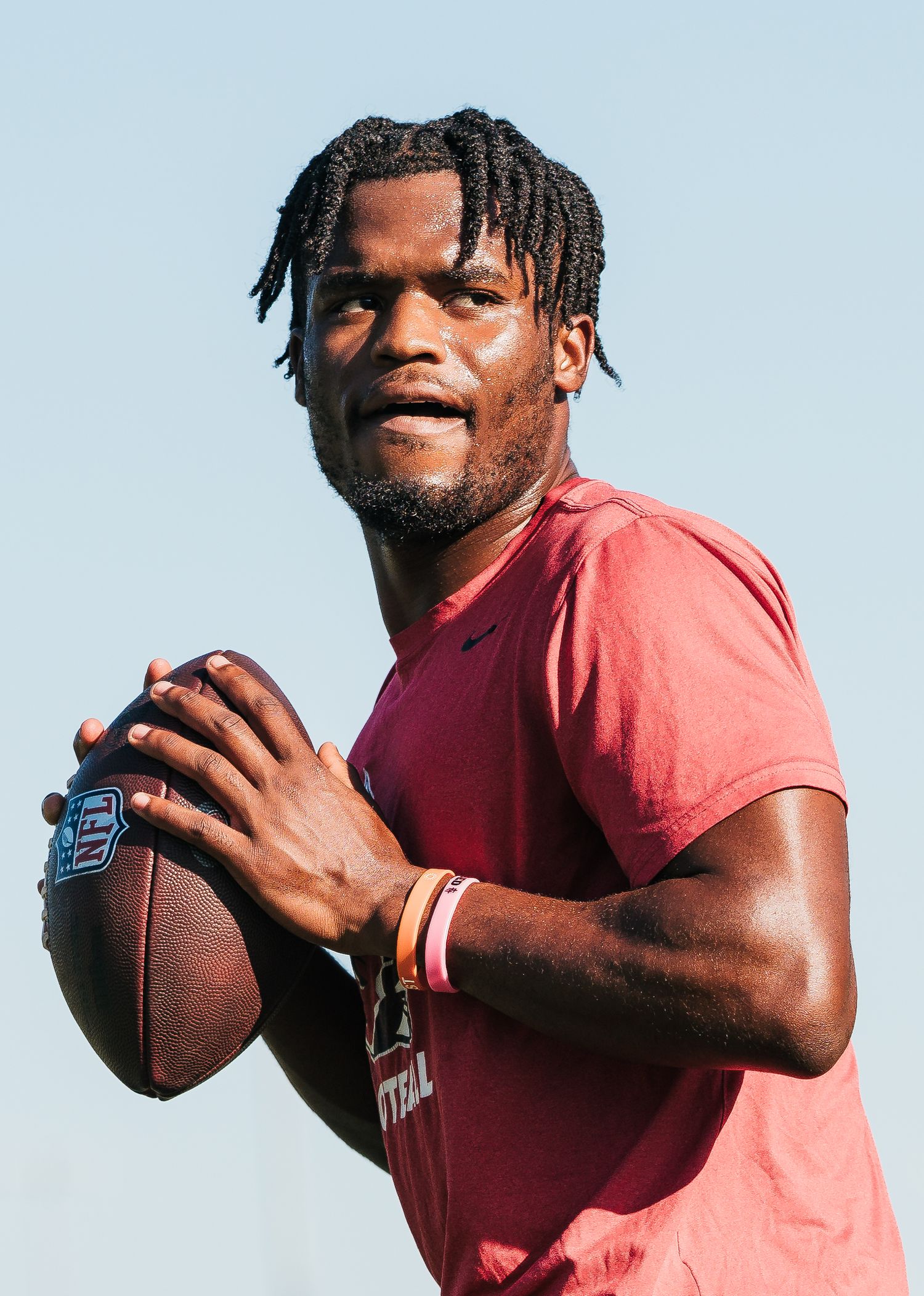 Young male athlete in red shirt holds football in throwing position outdoors on a clear day.