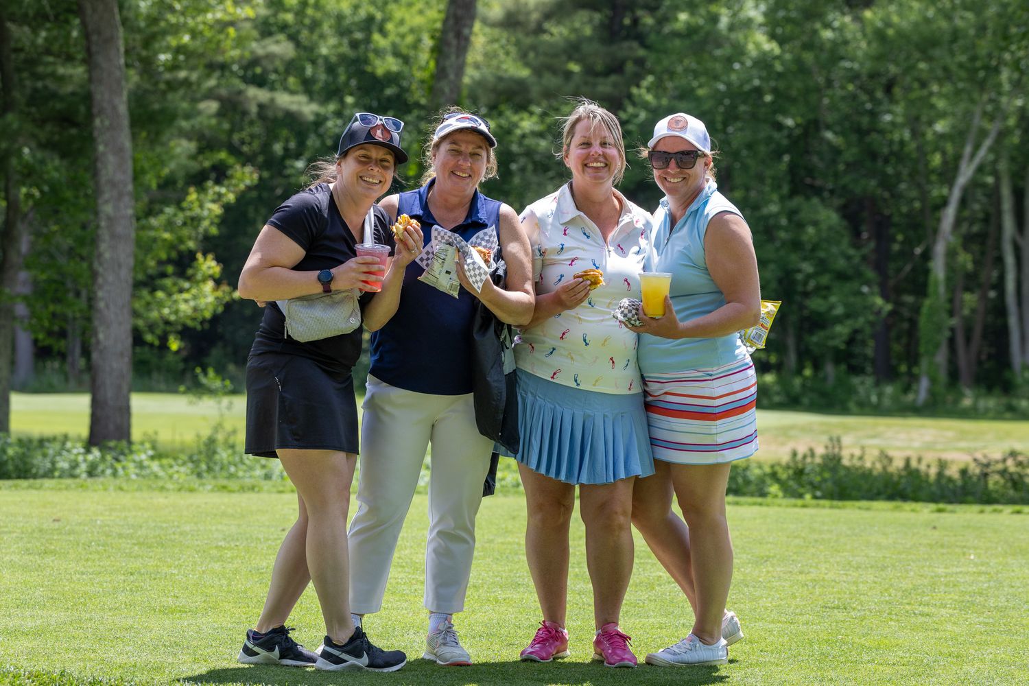 Four women golfers smiling and holding drinks on a sunny golf course surrounded by lush green trees.