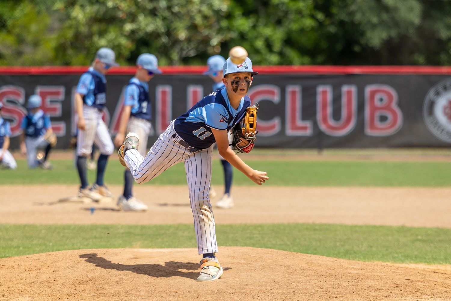 Youth baseball pitcher in blue uniform mid-throw on a sunny day at a baseball field.