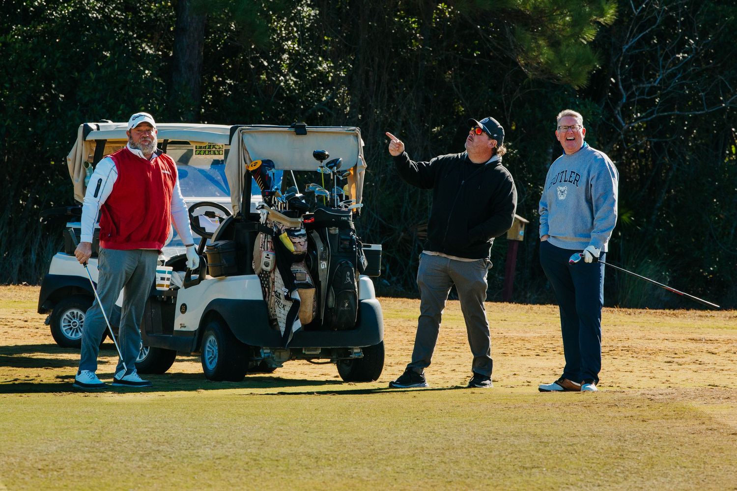 Three men standing near a golf cart on a sunny golf course, one pointing in the distance.