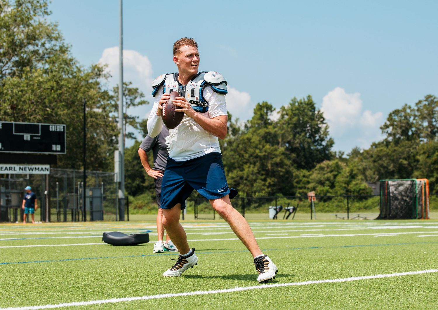 Football player in white uniform smiling while holding a ball during an outdoor practice session on a sunny day.