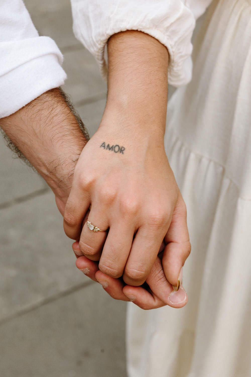 Close-up detail shot of hands showing tattoo and engagement ring against white fabric.