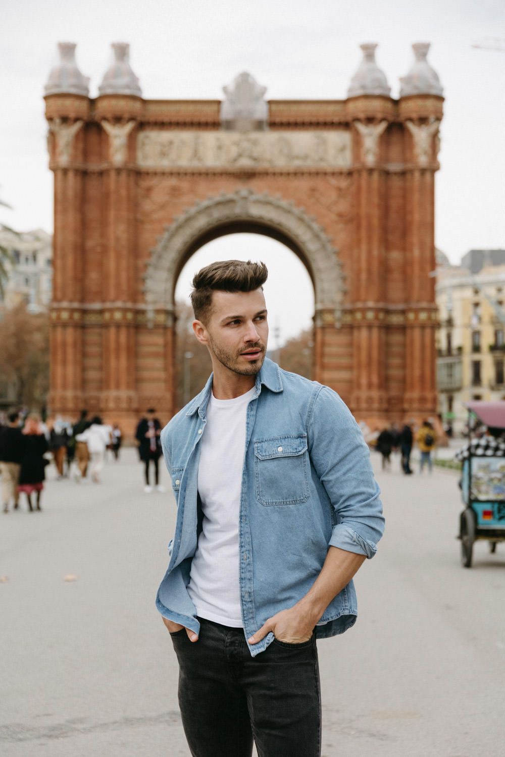 Man in denim jacket poses against historic Barcelona architecture during photoshoot.