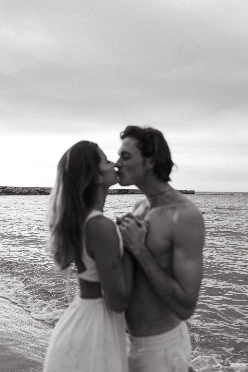 A couple shares romantic moments on a beach at sunset with ocean waves in background.
