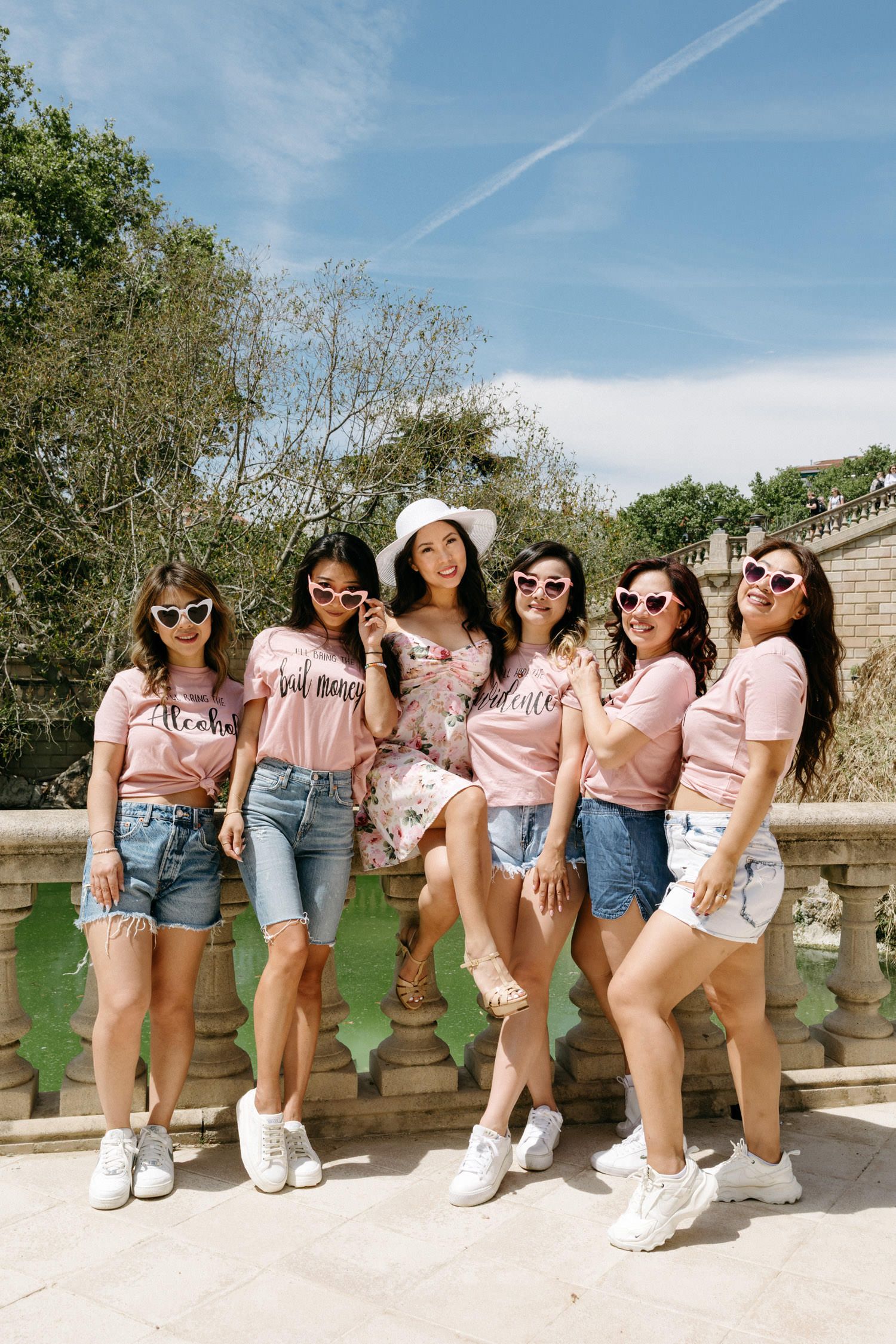 Group wearing matching pink shirts and denim pose on stone balustrade under blue sky.