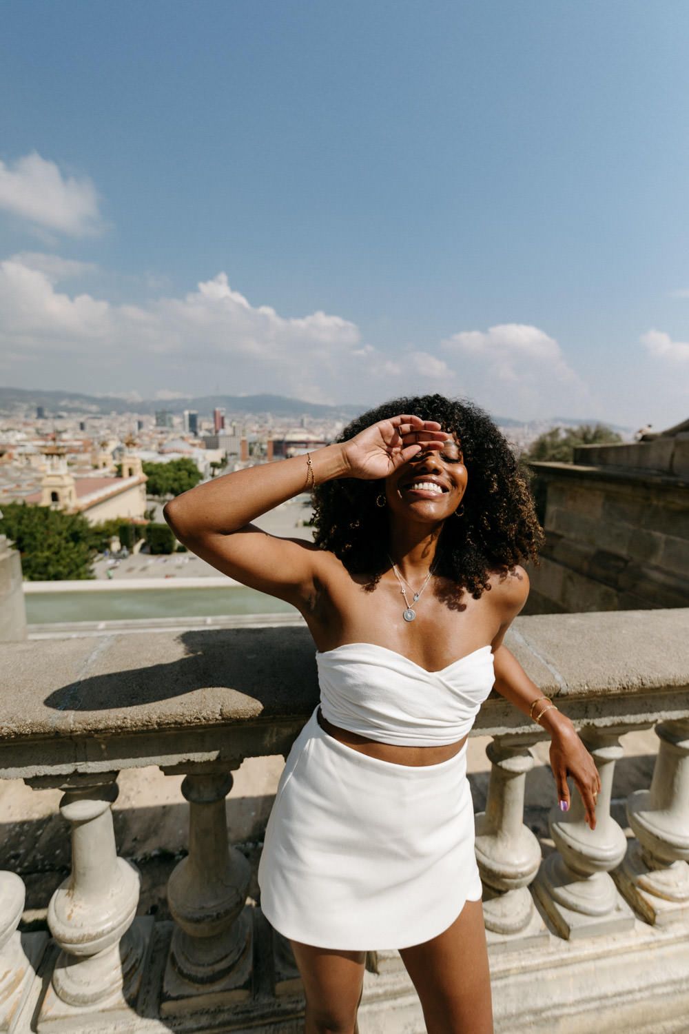 A person in white summer attire poses joyfully on a scenic rooftop terrace overlooking a Mediterranean cityscape.