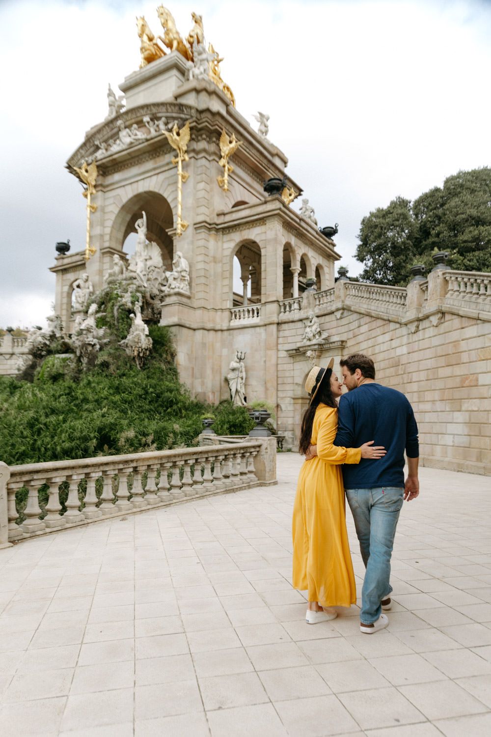 Couple strolling along ornate stone balustrade with golden statues at Barcelona's Parc de la Ciutadella.