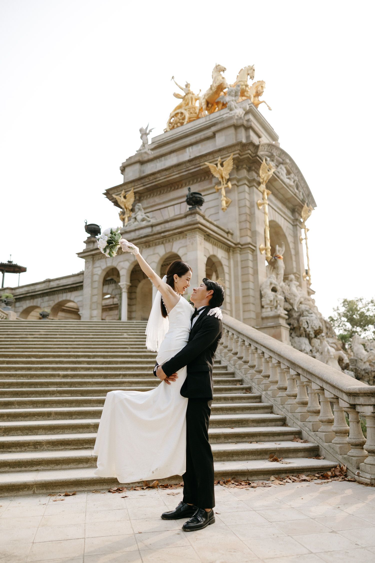 Wedding couple poses dramatically on grand ornate stone staircase with golden architectural details.