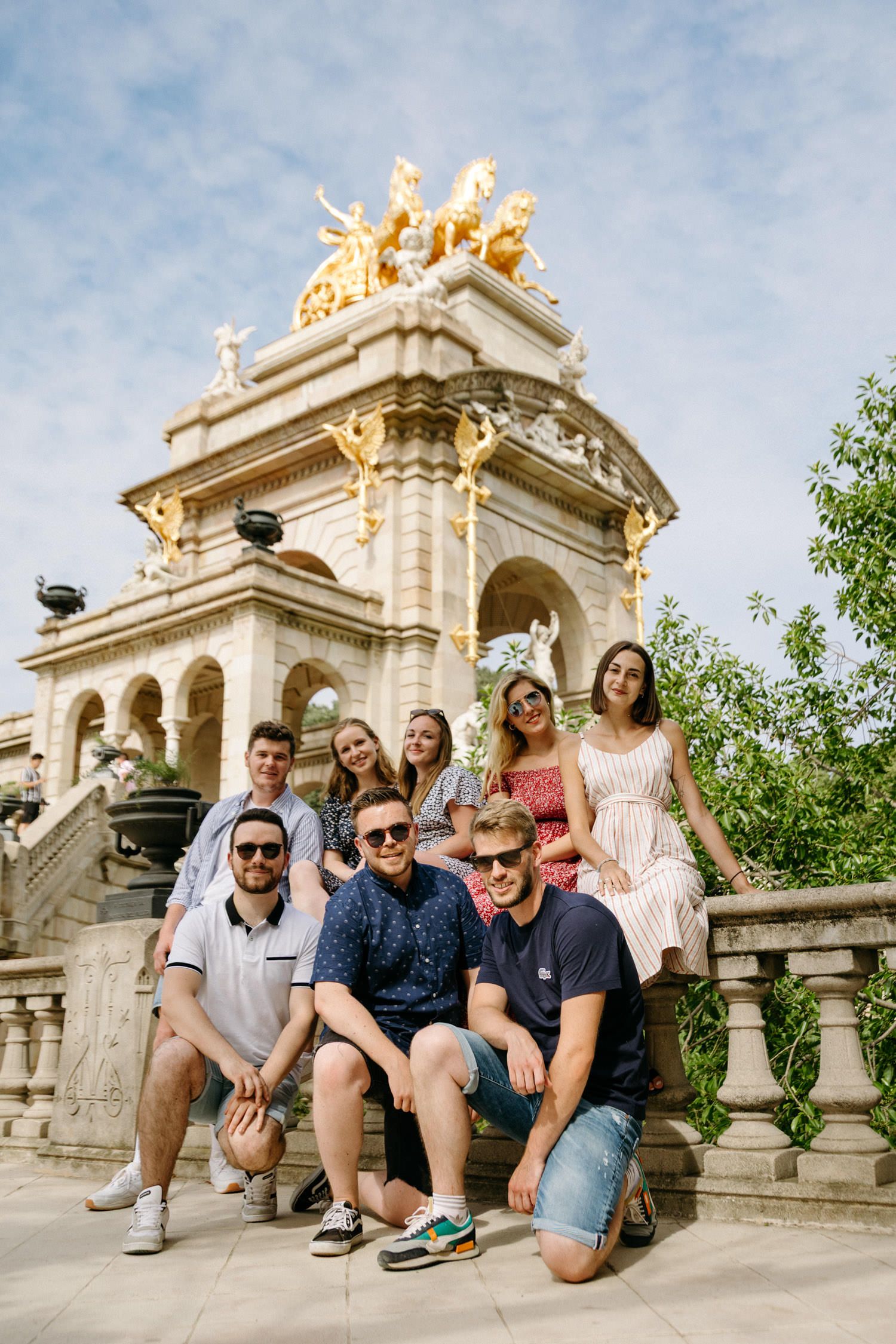 Group of friends sitting on ornate stone balustrade with decorative architecture and blue sky in background.