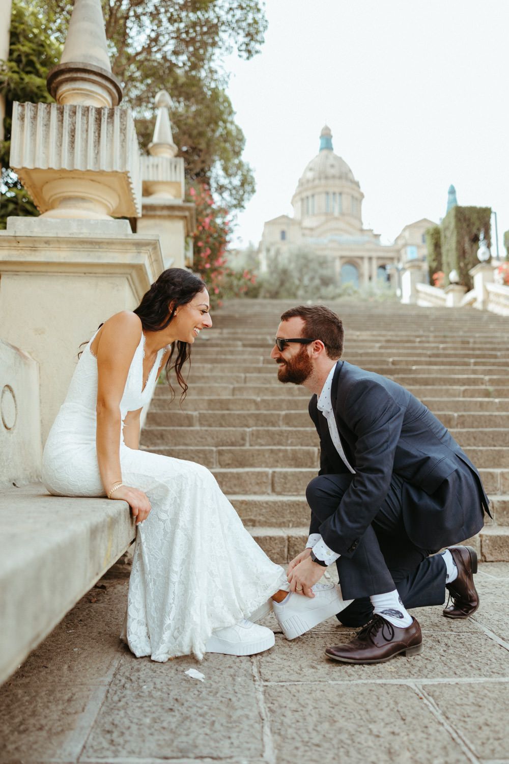 A couple in wedding attire playfully poses on stone steps with a domed building in the background.