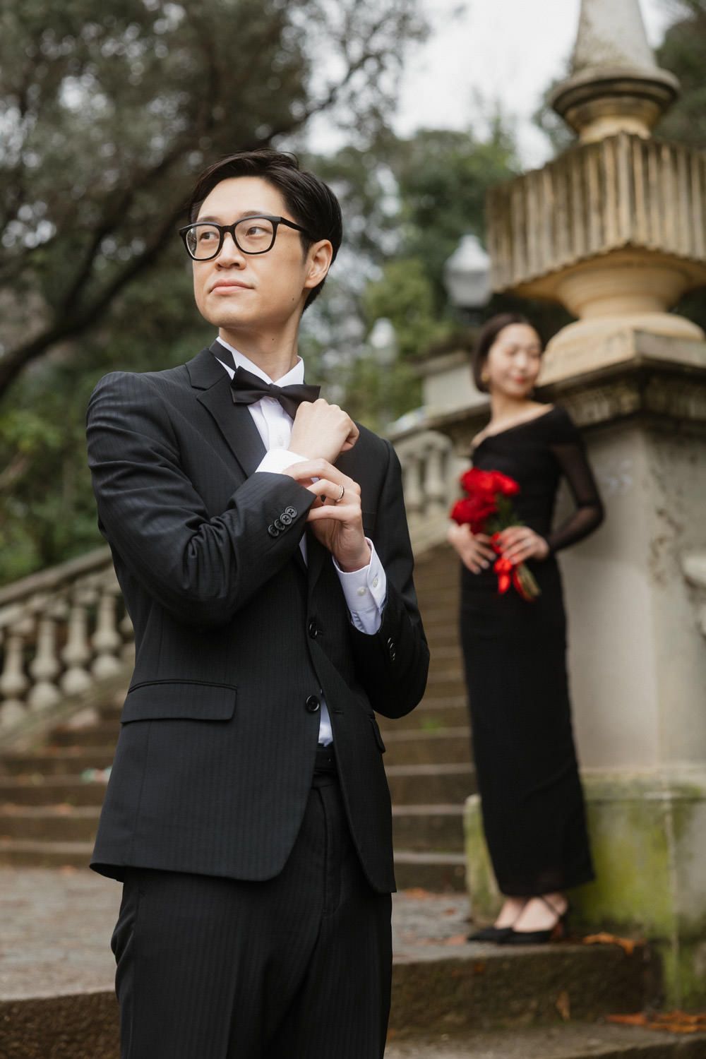 Elegant wedding couple poses on stone steps with groom in black tuxedo and bow tie while bride holds red bouquet in background.