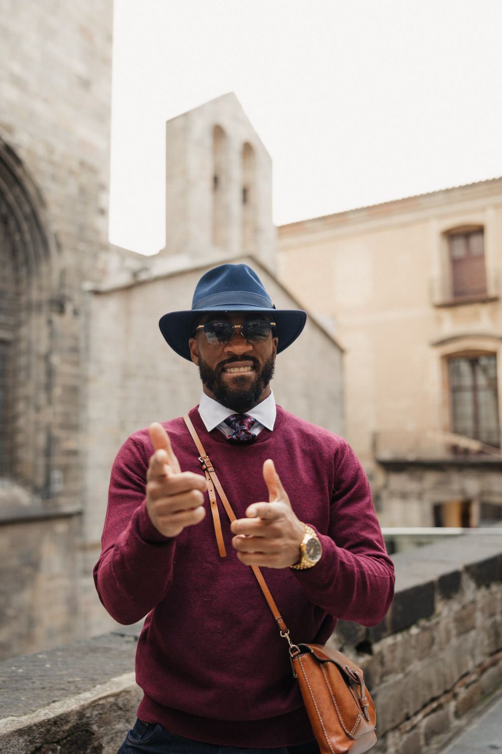 Person wearing a burgundy sweater and hat gestures enthusiastically while standing near historic stone buildings.