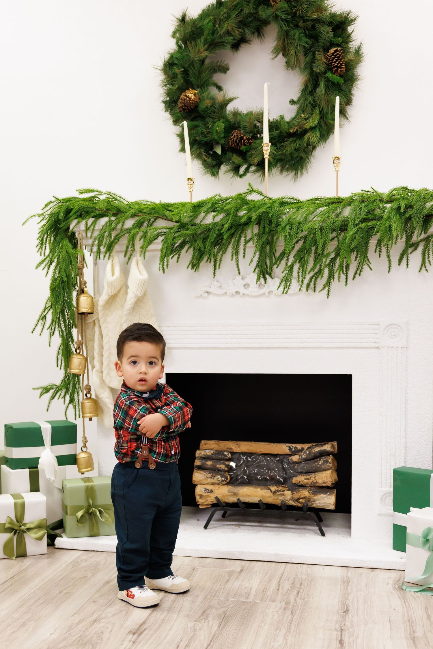 Toddler boy standing by a decorated fireplace with Christmas wreath, garland, and green gift boxes.