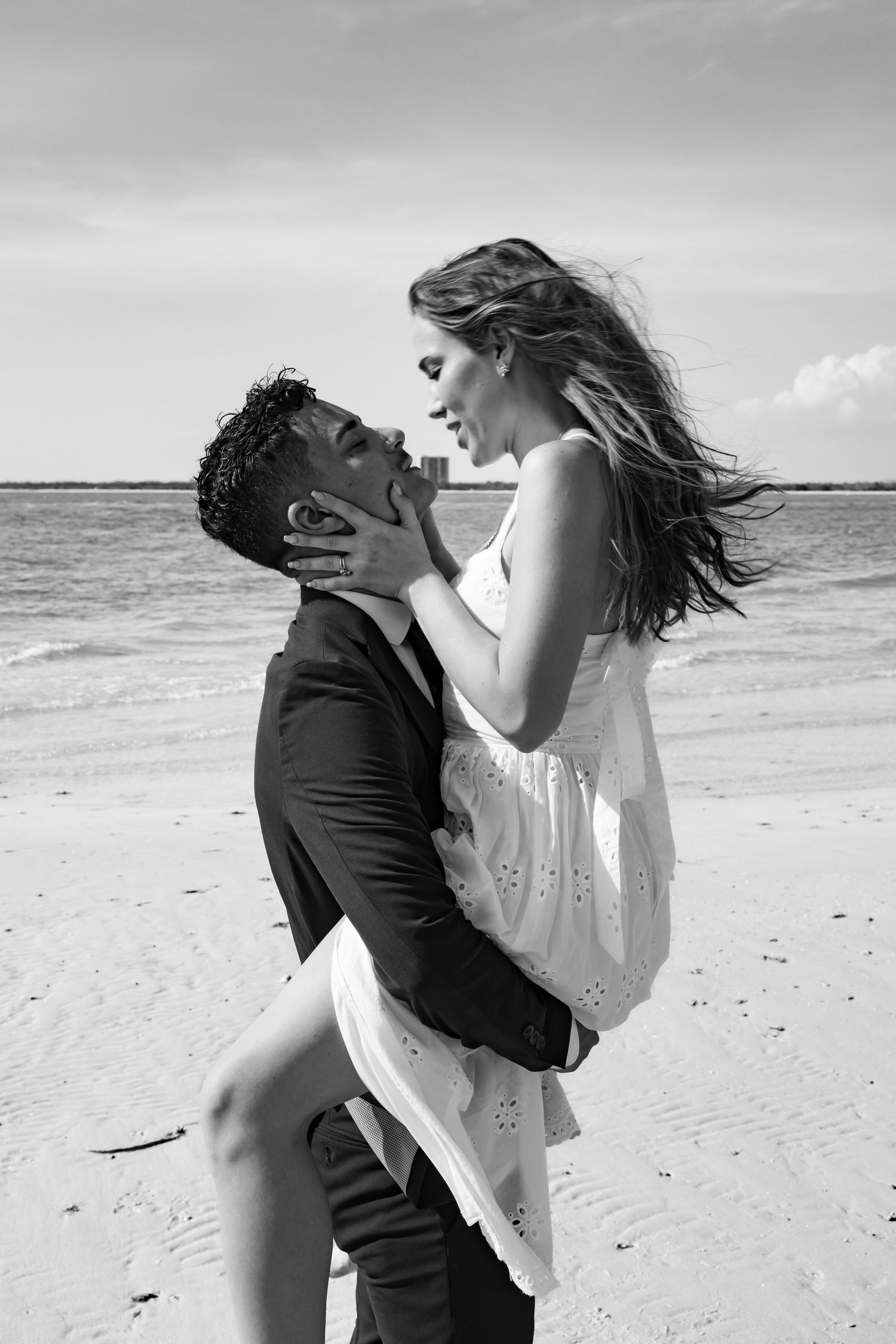 Black and white photo of a romantic couple embracing on a beach, man lifting woman in white dress near ocean waves.