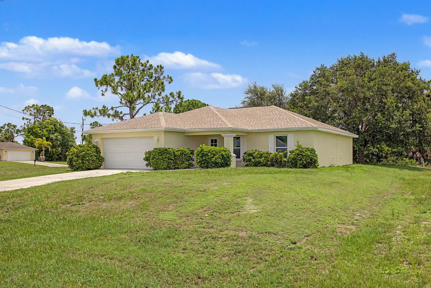 Single-story beige stucco home with a tile roof, attached garage, and a landscaped green lawn under a blue sky with scattered clouds.