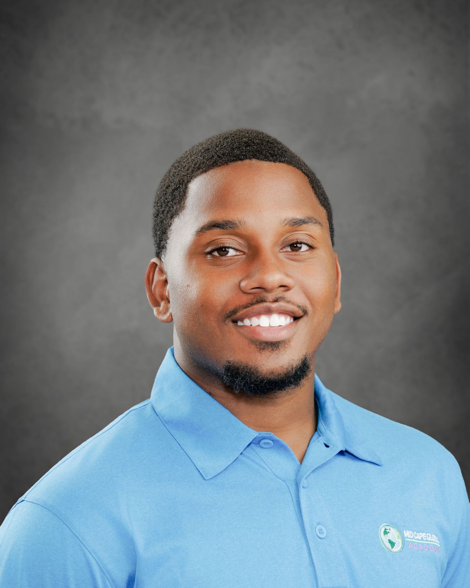 Smiling man with short dark hair and a goatee wearing a light blue polo shirt with a company logo against a gray background.
