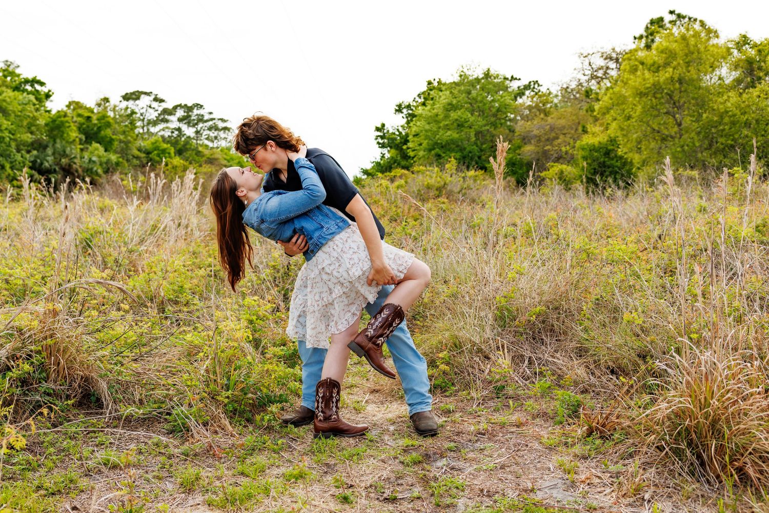 Couple sharing a romantic kiss in a field, man lifting woman in a loving embrace surrounded by lush greenery.