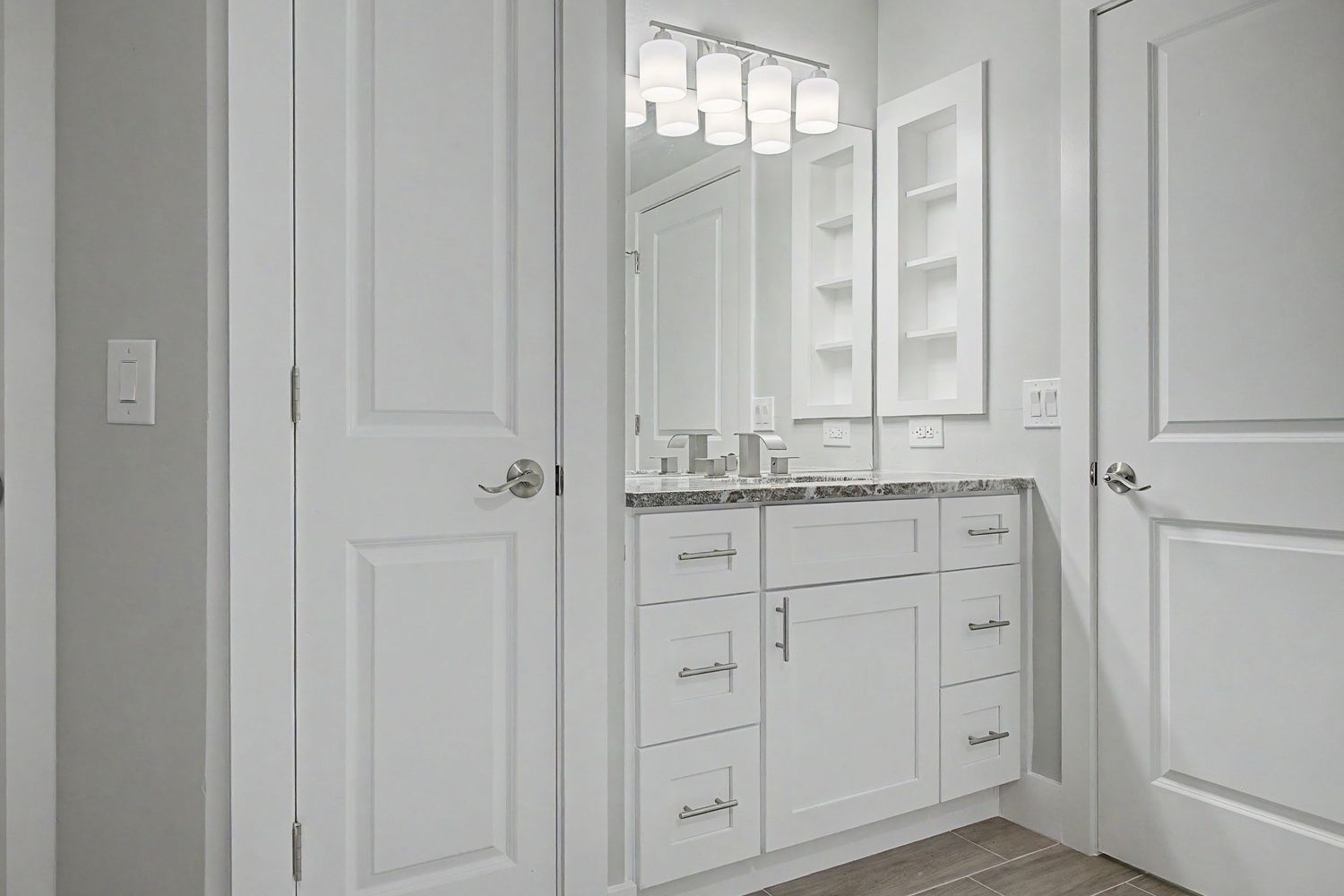 Bright bathroom with white shaker vanity cabinets, granite countertop, large mirror, recessed wall shelves, and wood-look flooring.