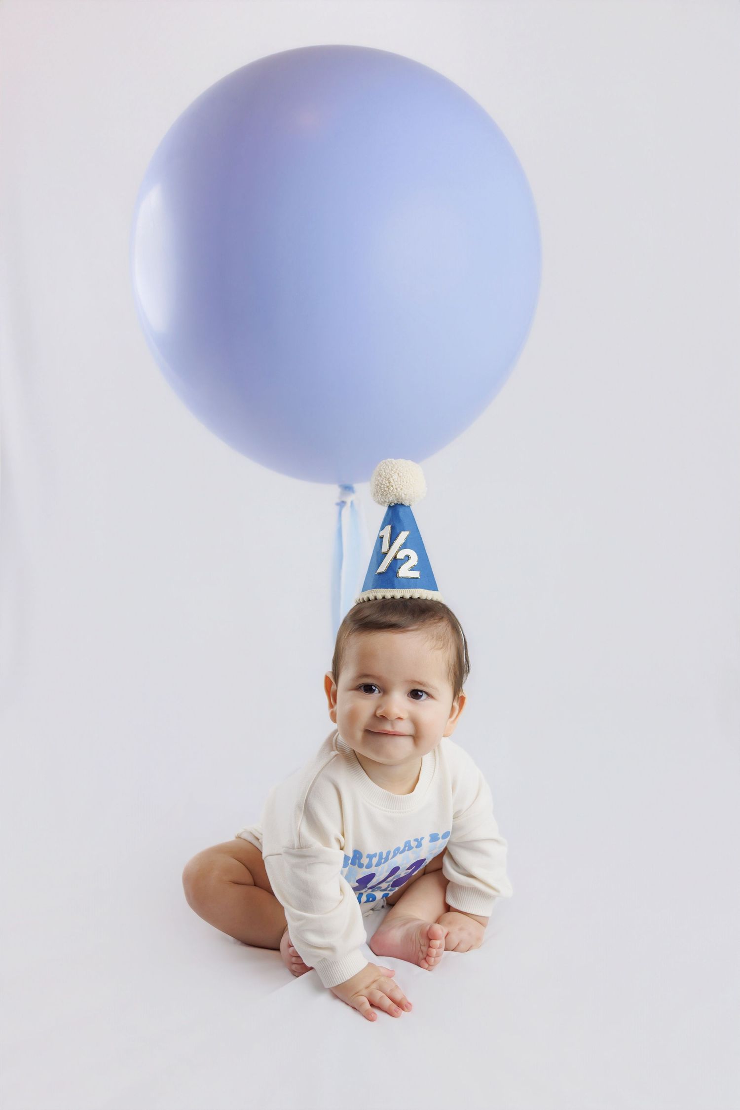Sweet baby in a half birthday hat sitting next to a large lavender balloon on a white background.