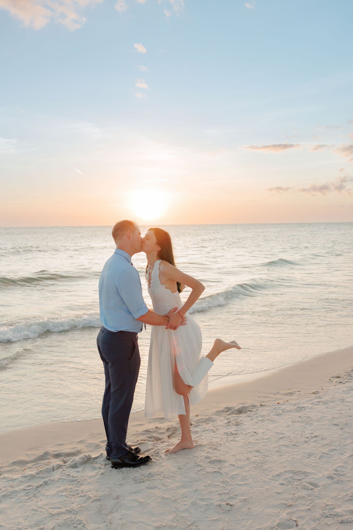 Couple sharing a romantic kiss on a white sand beach at sunset, woman in white dress with one leg raised.