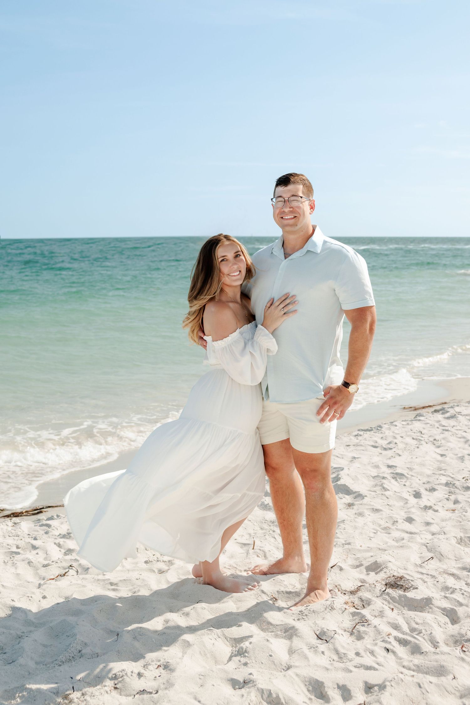 Couple in white outfits embracing on a sandy beach with turquoise ocean waves in the background.
