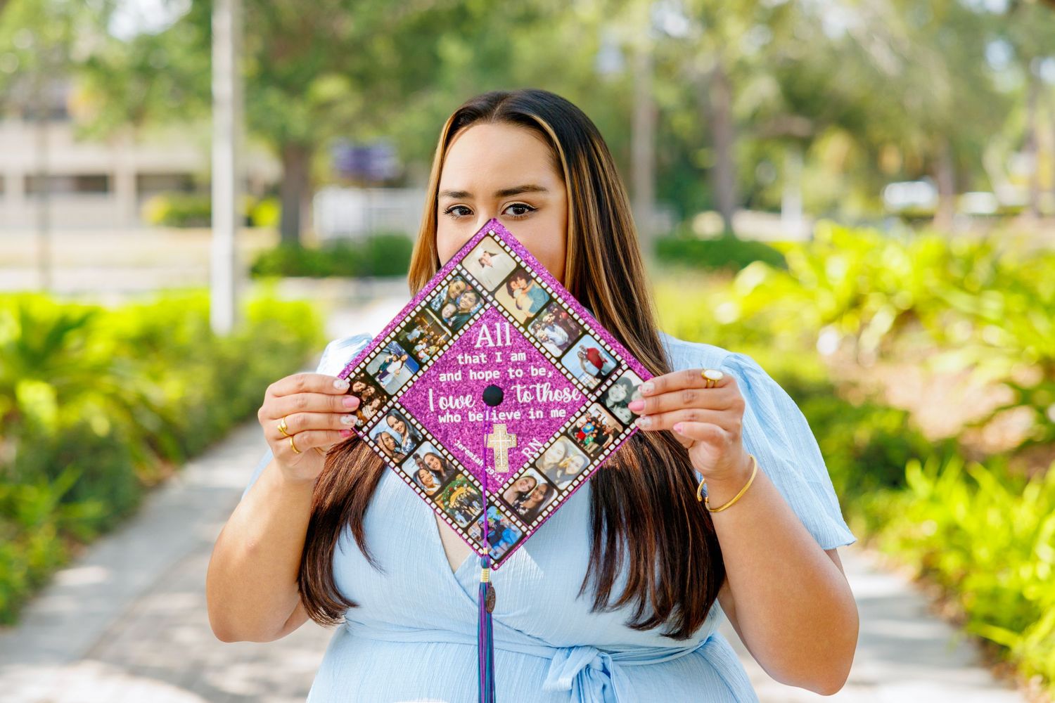 Graduate in a light blue dress holds up a decorated pink graduation cap covered with photos and handwritten text outdoors near green trees.