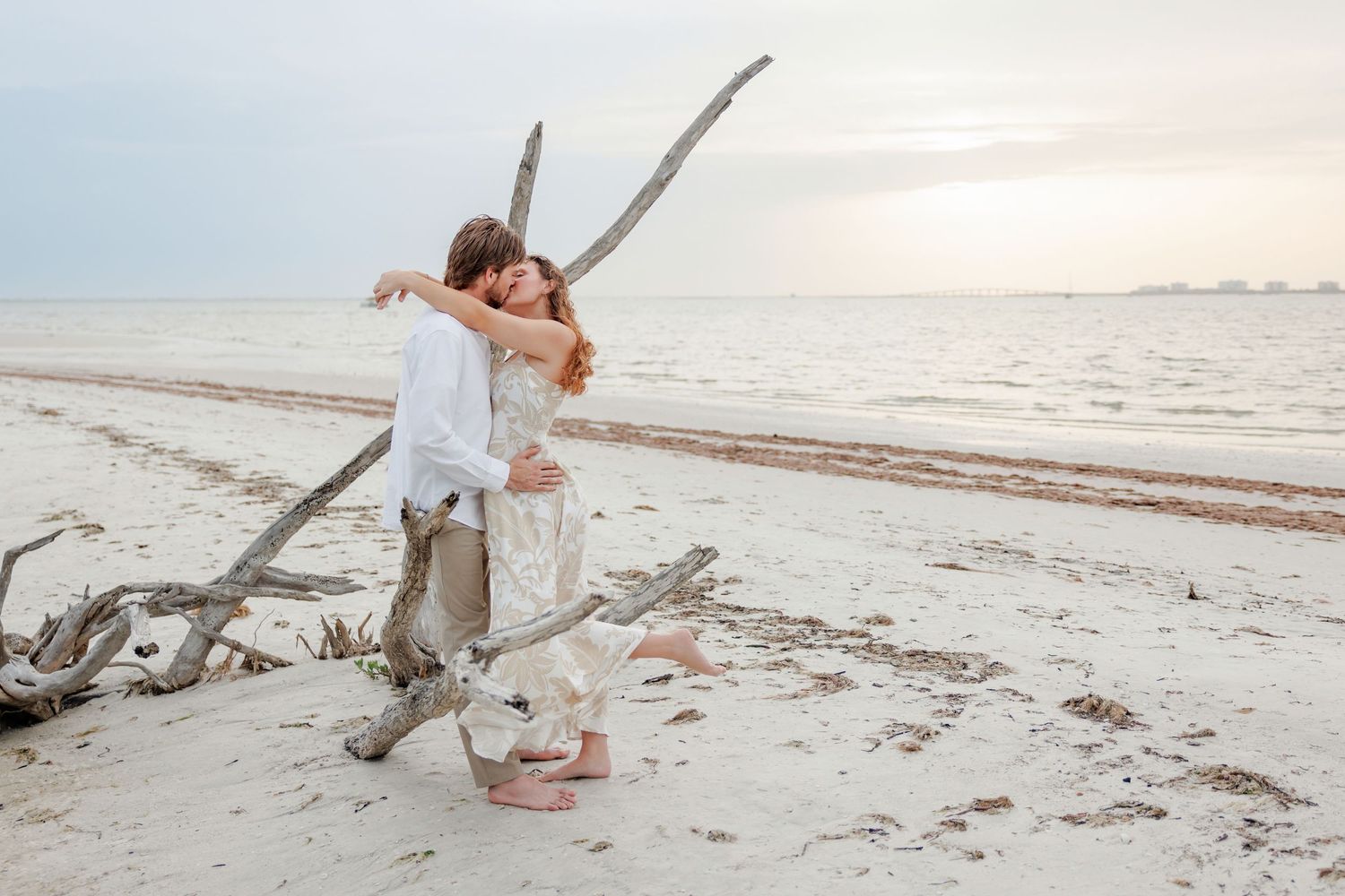 Couple embracing on a sandy beach beside driftwood at sunset, with calm ocean water and a soft cloudy sky in the background.