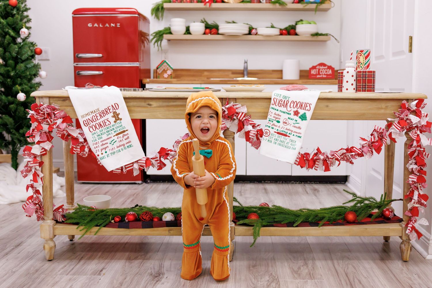 Toddler in gingerbread costume poses in festive Christmas kitchen with holiday decorations and signs.
