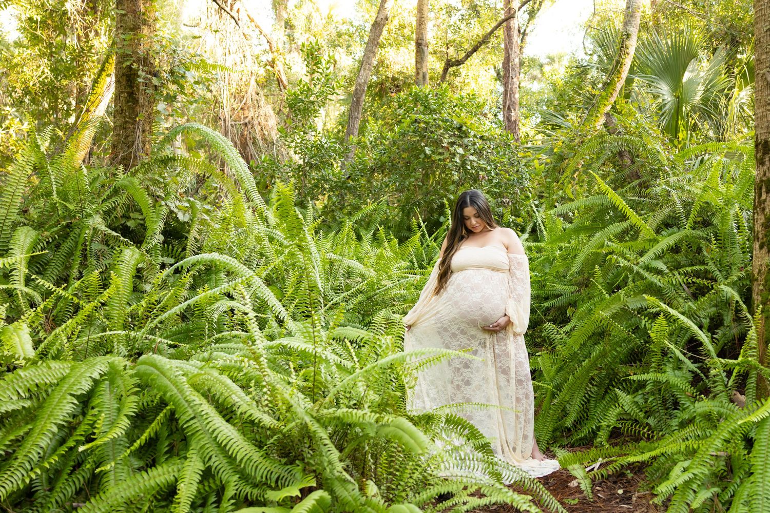 Pregnant woman in floral dress cradling baby bump surrounded by lush green ferns in a forest setting.