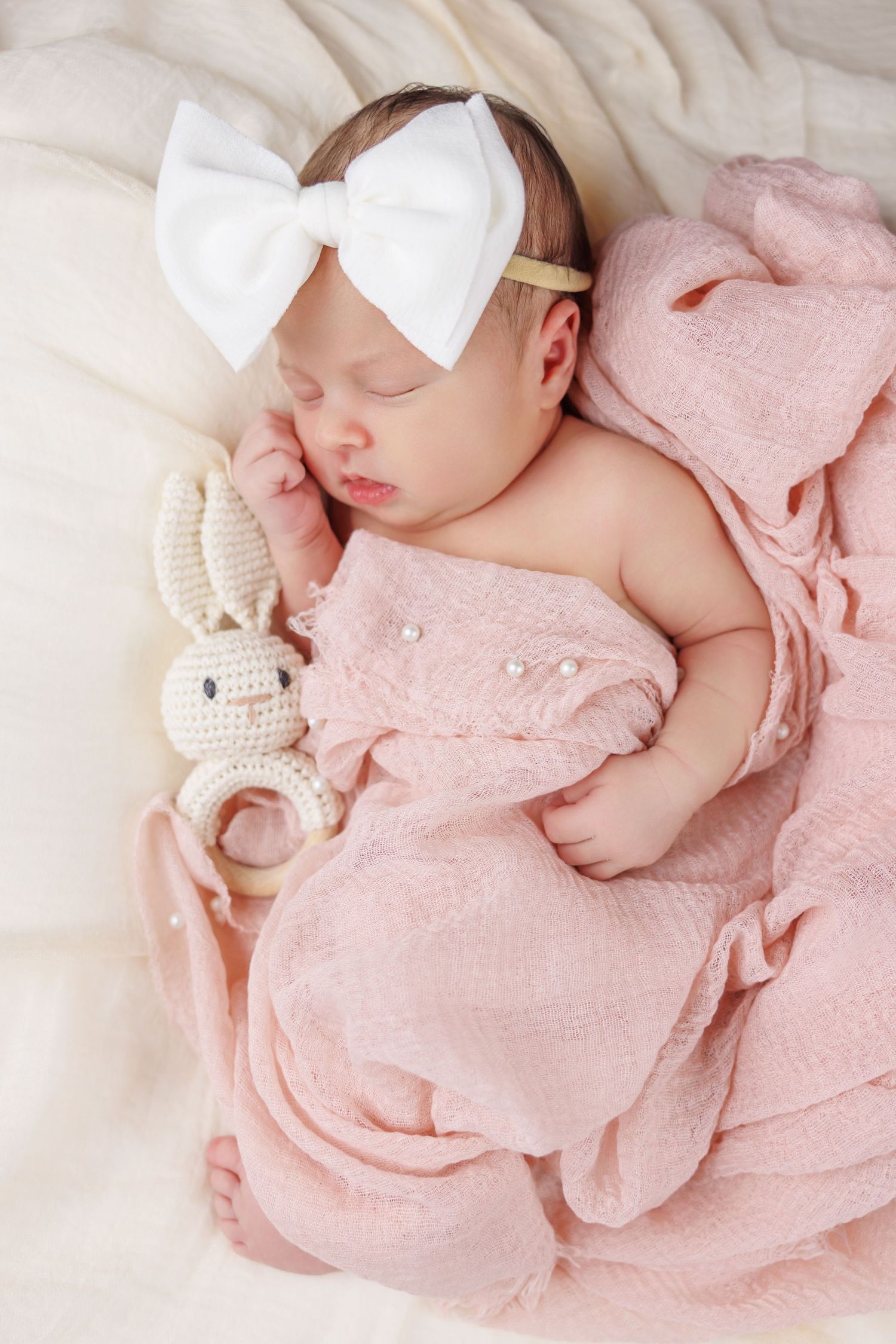 Newborn baby girl sleeping peacefully wrapped in pink muslin blanket with white bow headband and crochet bunny toy.