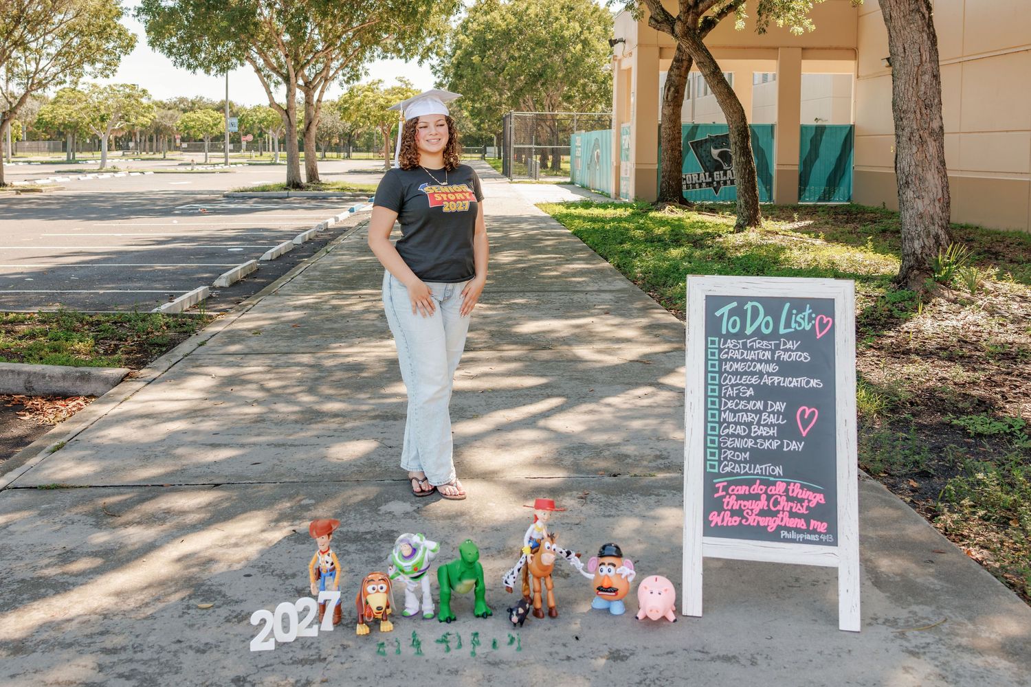 Woman in a gray graphic tee and white pants stands on a sunny sidewalk beside a chalkboard to-do list sign and toy figures marked 2021.