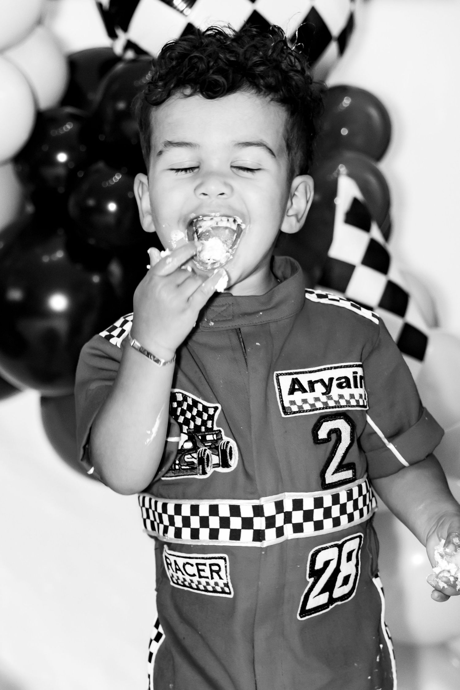 Boy in racing outfit eating birthday cake smash, balloons and checkered flags in background.