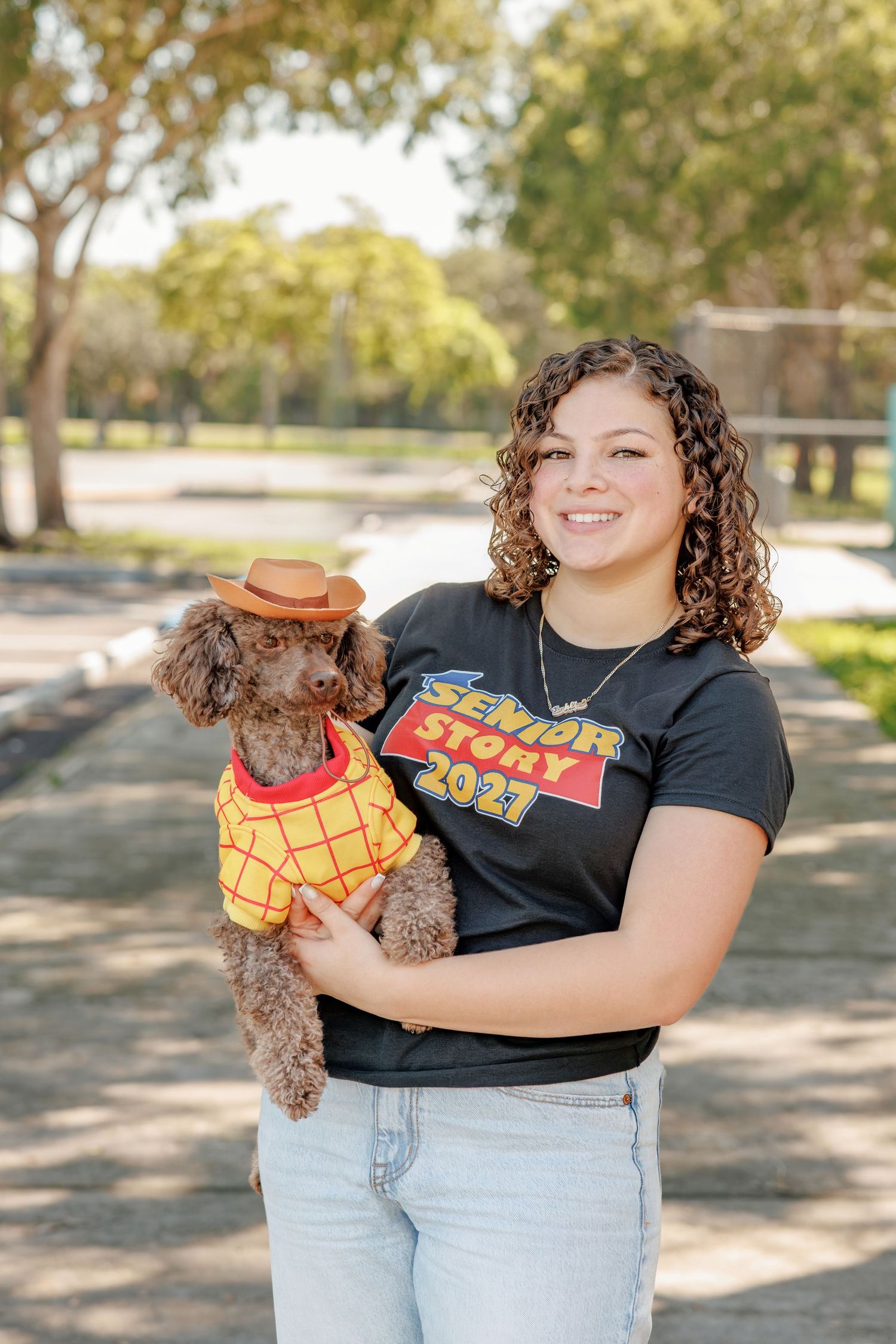 Smiling young woman with curly hair in a Senior Story 2021 shirt holds a brown poodle wearing a cowboy hat and yellow plaid vest outdoors.