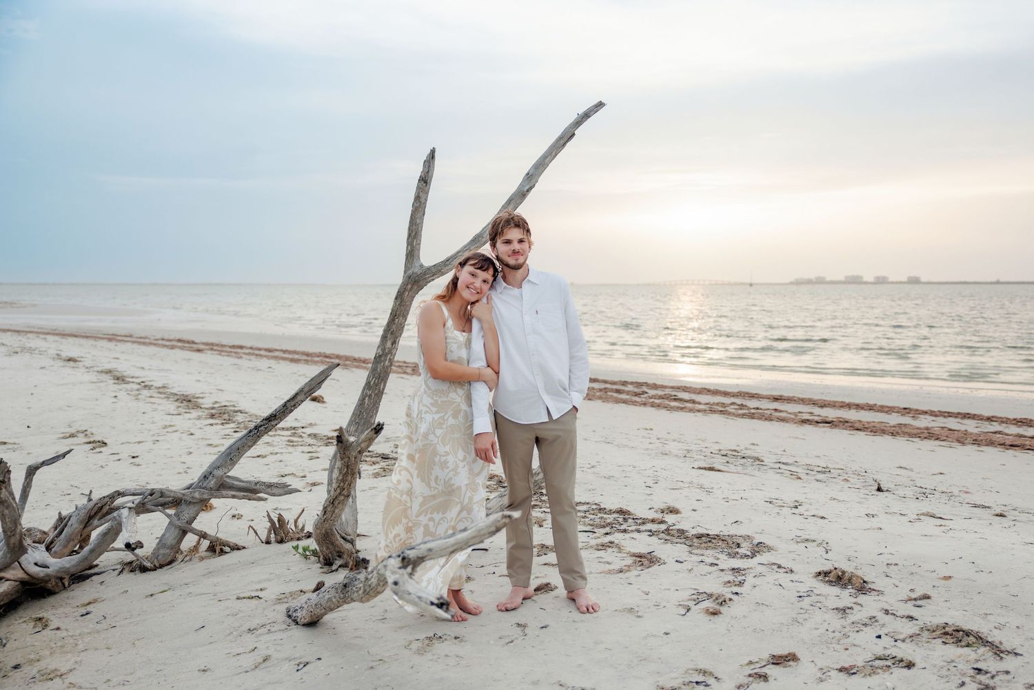 Barefoot couple embracing beside weathered driftwood on a sandy beach at sunset, with calm ocean waters and a soft hazy sky behind them.