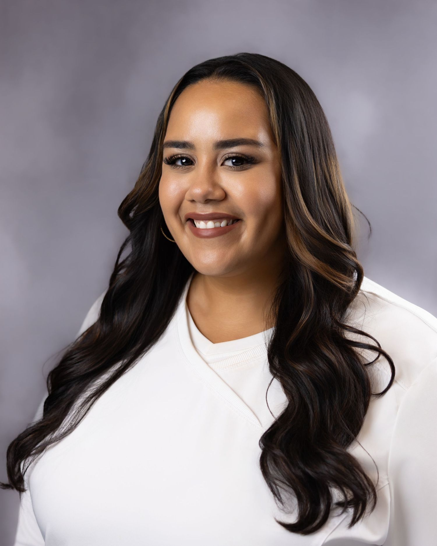 Young woman with long dark wavy hair smiling in a white top against a gray studio background.