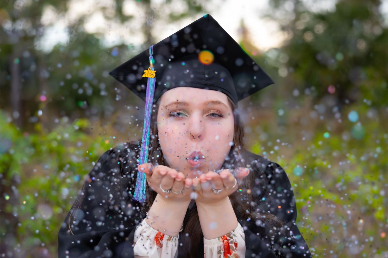 Graduate in cap and gown blowing colorful glitter confetti from her hands outdoors.