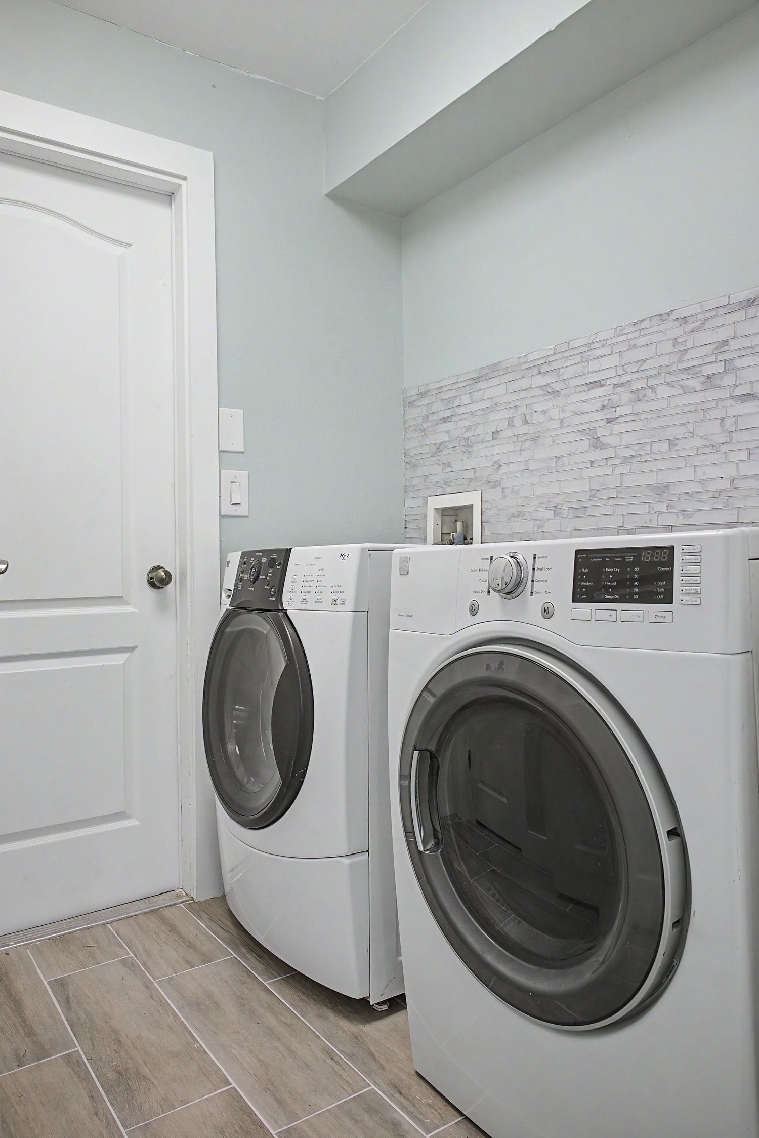 Laundry room with front-load white washer and dryer, light blue walls, a stone tile backsplash and wood-look tile flooring.